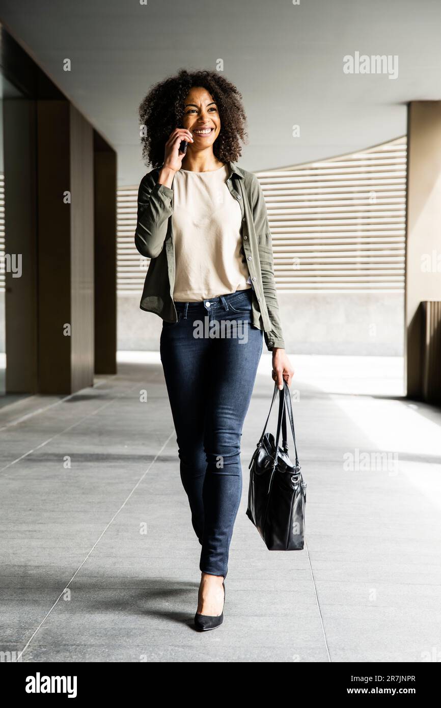 Donna sorridente che cammina in un edificio utilizzando un telefono che porta una borsa. Foto Stock