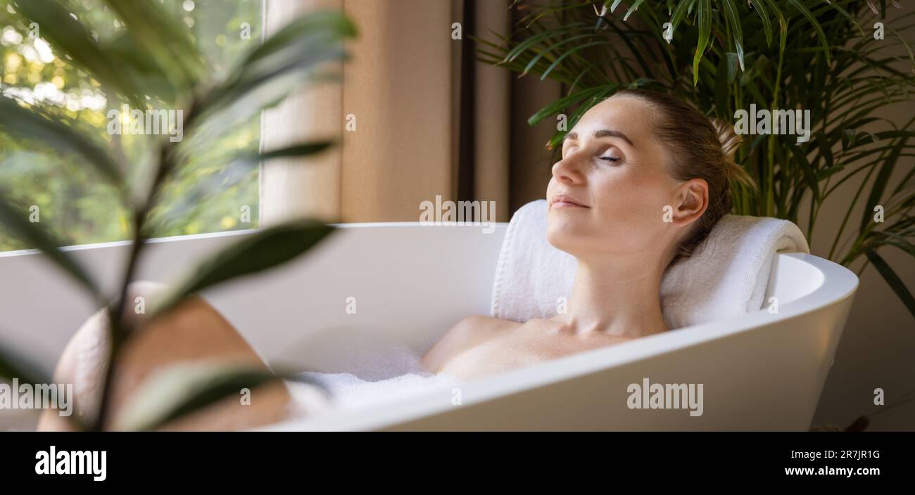 donna che si rilassa nella vasca da bagno con gli occhi chiusi nel bagno con piante tropicali verdi. trattamento termale, cura del corpo e della pelle. banner con spazio di copia Foto Stock