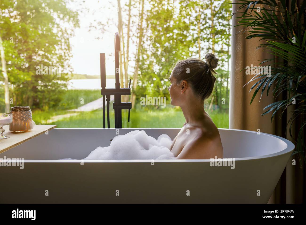 giovane donna che si rilassa nella vasca da bagno piena di schiuma e che guarda fuori dalla finestra del bagno di casa Foto Stock