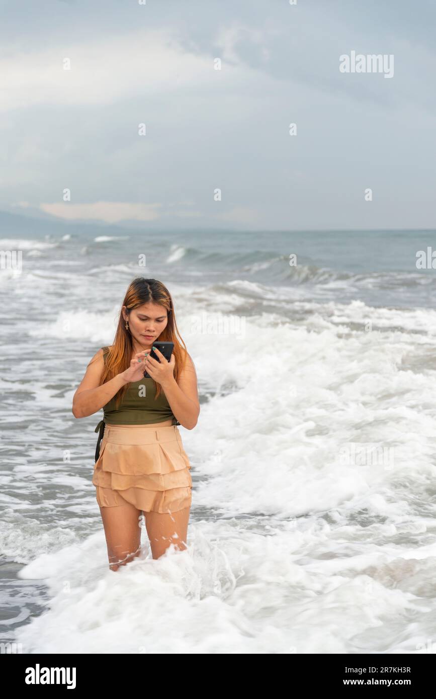 Tenendo il suo telefono per selfie, su una spiaggia vicino Manila al tramonto, aimlessly passeggiando le calde acque costiere, lungo le sabbie lisce, avendo divertimento, ballando aroun Foto Stock