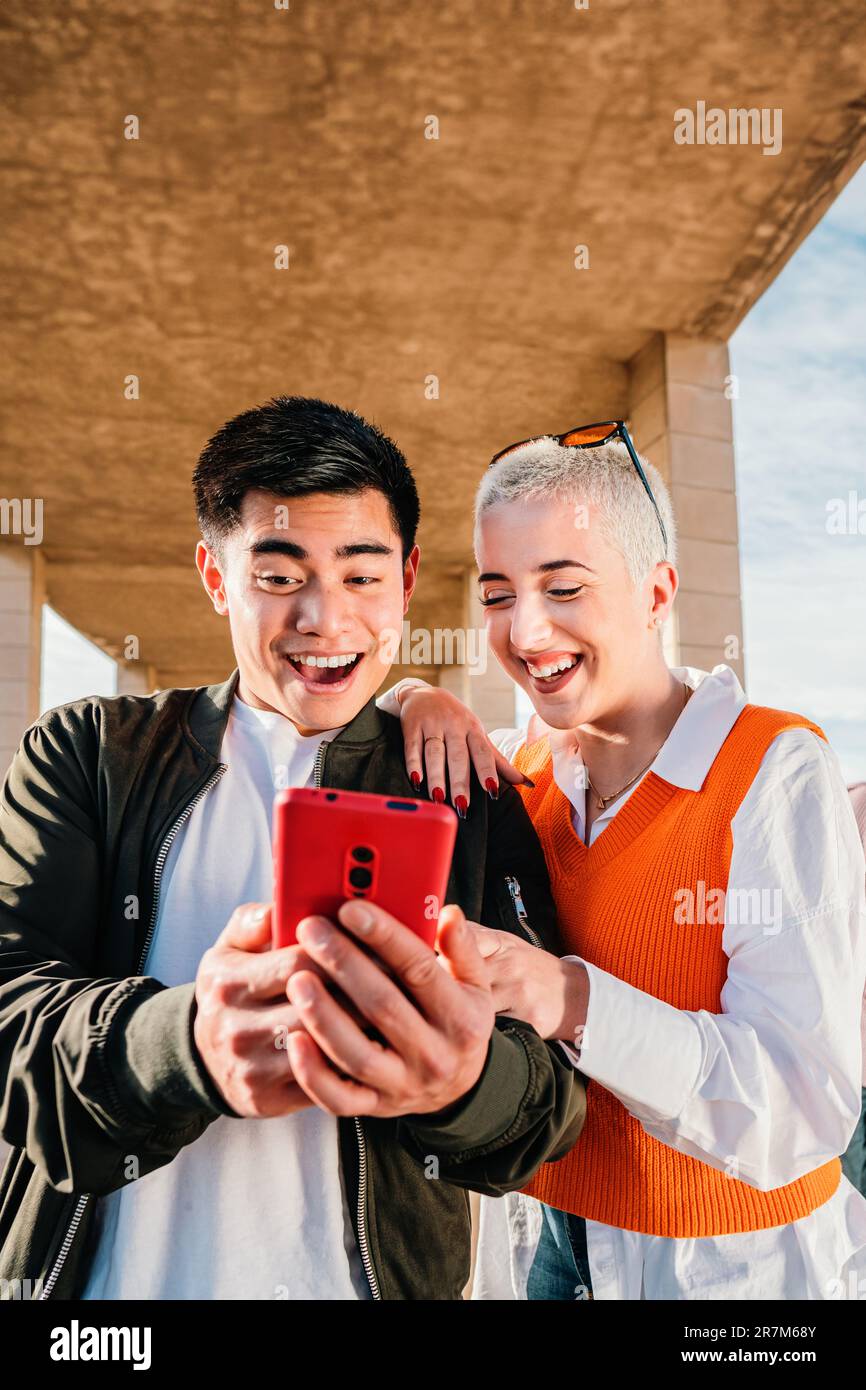 Ritratto verticale di una giovane coppia utilizzando un cellulare per guardare le foto dell'app social media. Persone multirazziali in possesso di un telefono cellulare e divertirsi insieme. Ragazzo e ragazza con smartphone. Foto di alta qualità Foto Stock