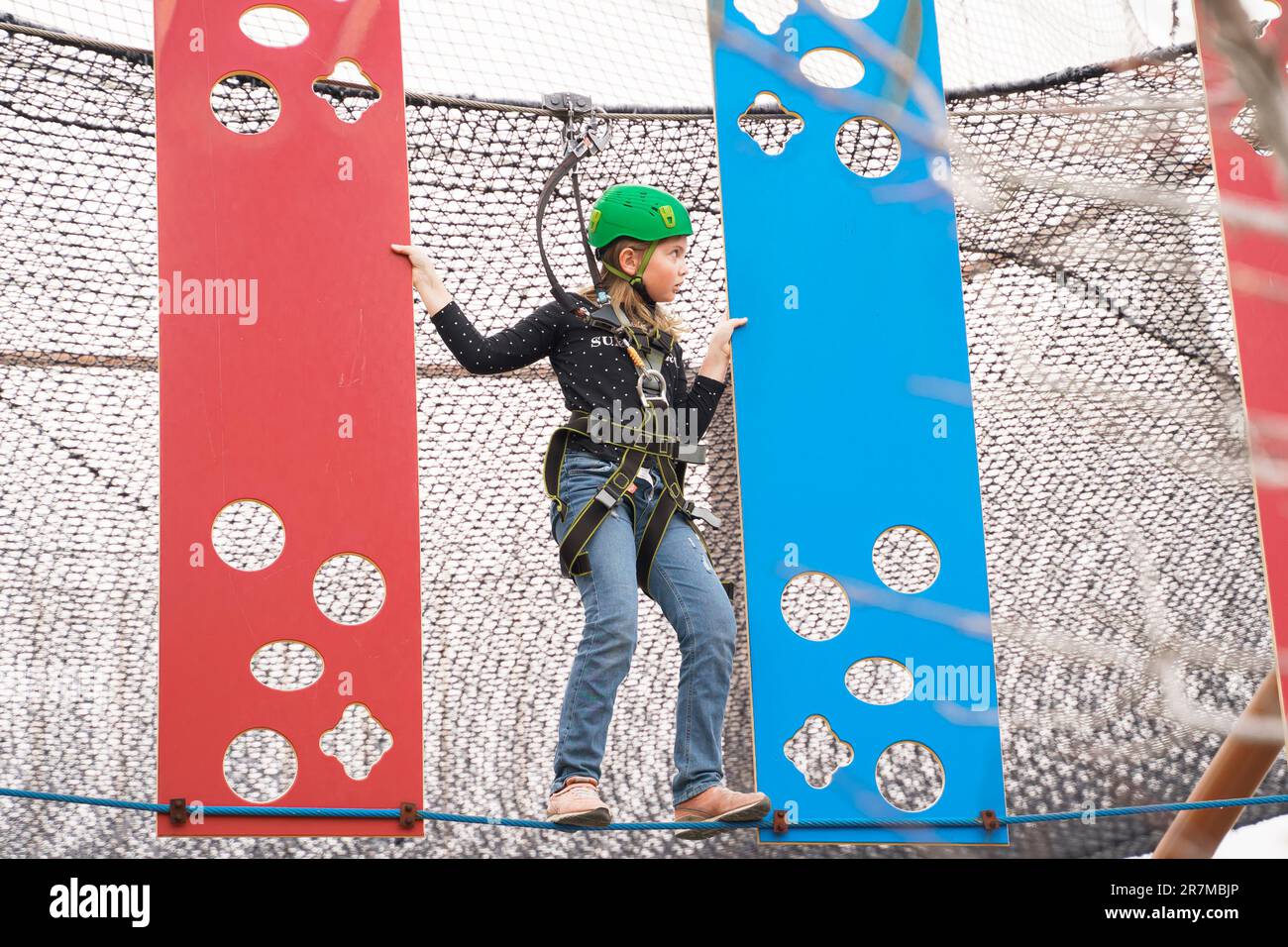 Adolescente ragazza in attrezzatura di arrampicata imbracatura, casco verde sport di sicurezza. Parco divertimenti Rope. Fissaggio del moschettone alla fune di sicurezza. Hangin Foto Stock