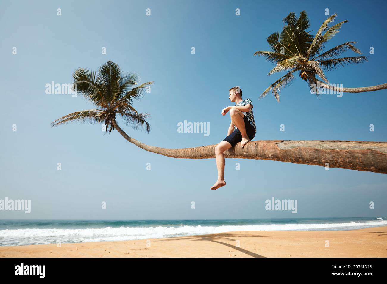 Uomo seduto sul tronco di palma e con vista sul mare. Spiaggia di sabbia idilliica in Sri Lanka. Foto Stock