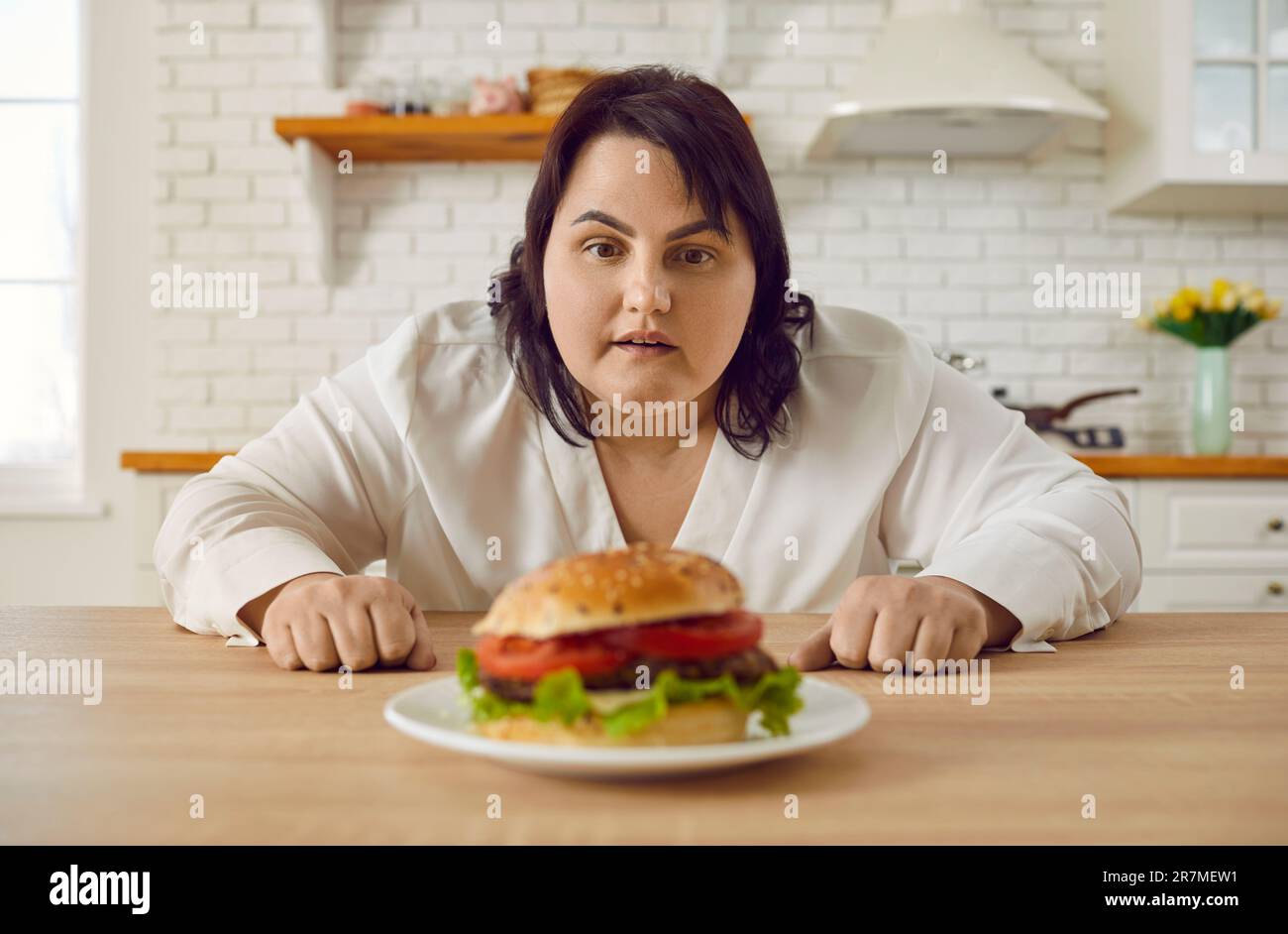 Giovane donna grassa eccitata seduta al tavolo in cucina guardando il piatto con grande hamburger. Foto Stock