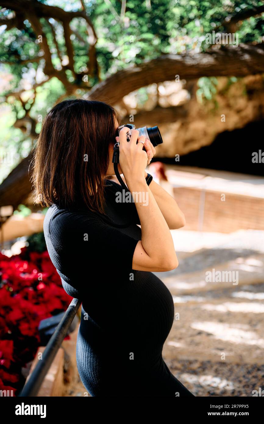 ritratto di una giovane donna in un abito stretto nei suoi primi mesi di gravidanza scattando foto con una fotocamera antica e guardando la fotocamera. Foto Stock