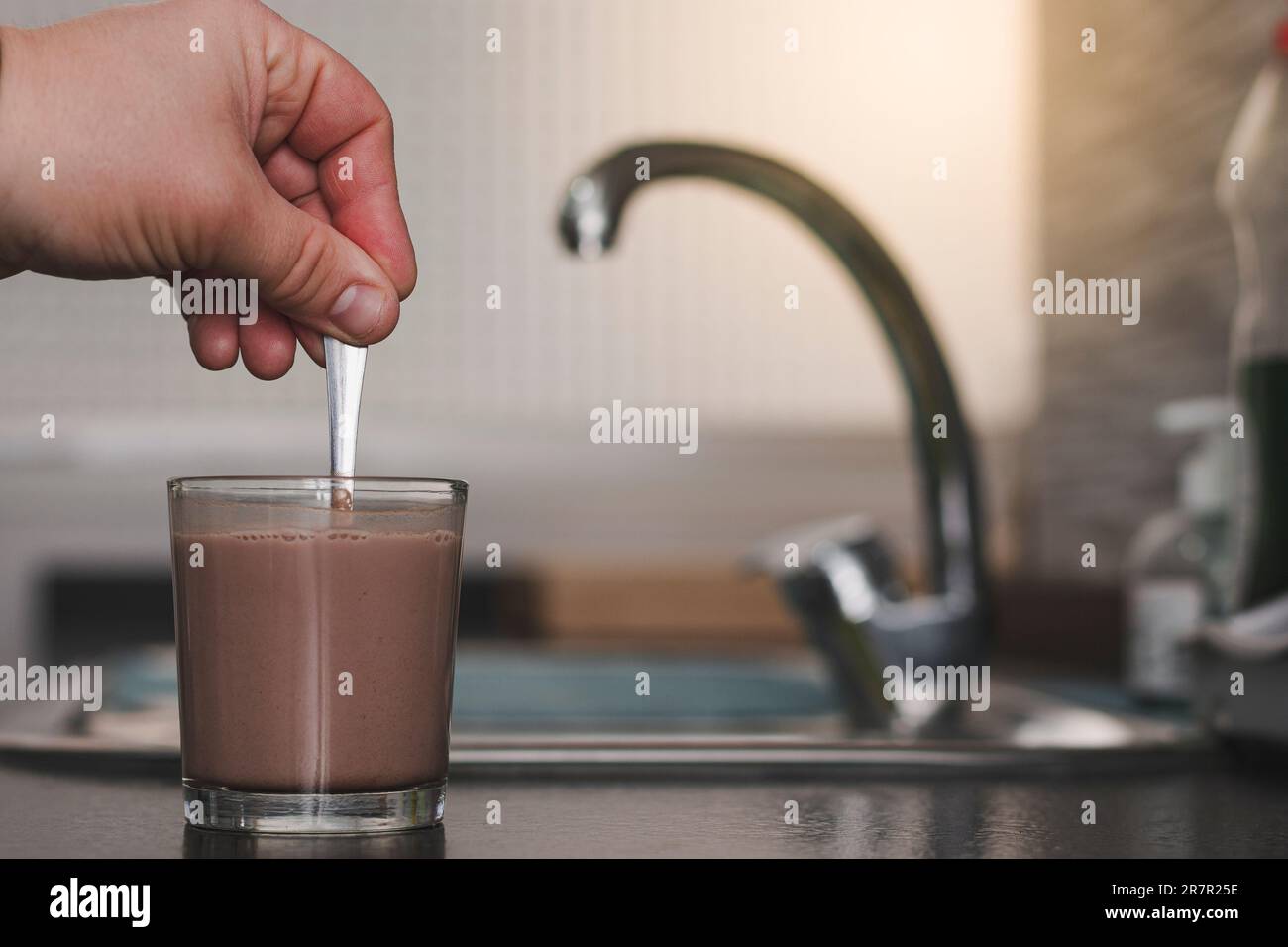 Primo piano di una mano maschio adulta caucasica irriconoscibile che mescola latte e cacao in un bicchiere con un cucchiaio su una panca da cucina. Foto Stock