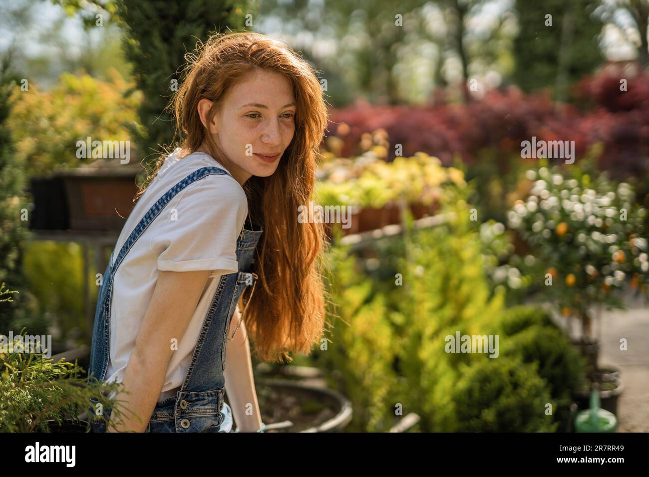 Una foto di una botanica femminile rossa seduta in giardino e guardando qualcosa di bello con un lieve sorriso sul viso Foto Stock