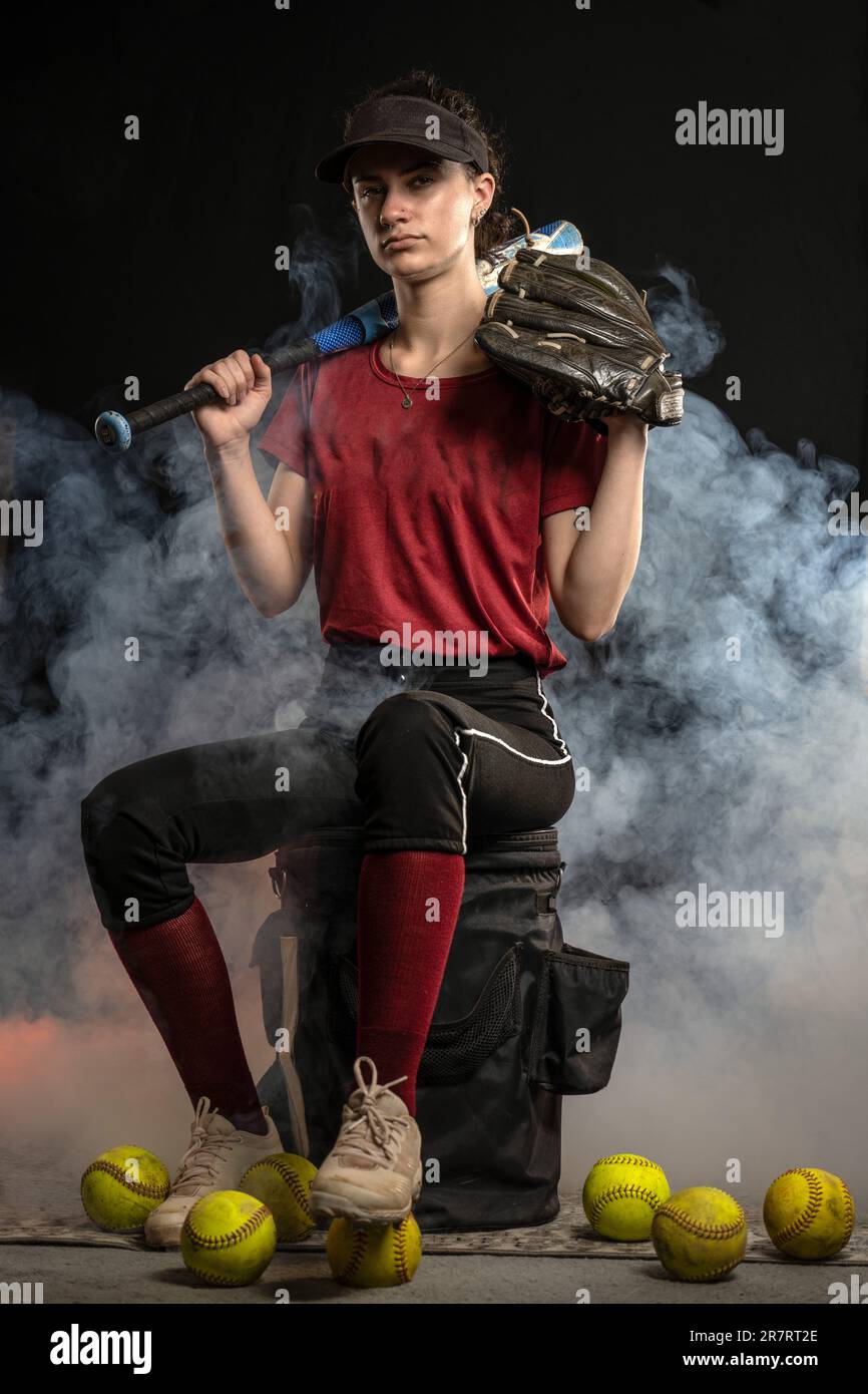 Il giocatore di softball sportivo a pitch veloce in uniforme rossa e nera si siede sul secchio degli allenatori con sfondo appannato. Foto Stock