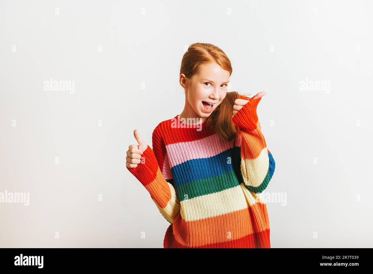 Studio shot di bella ragazza adolescente con i pollici grandi in su, indossando pullover colorato, posando su sfondo bianco Foto Stock