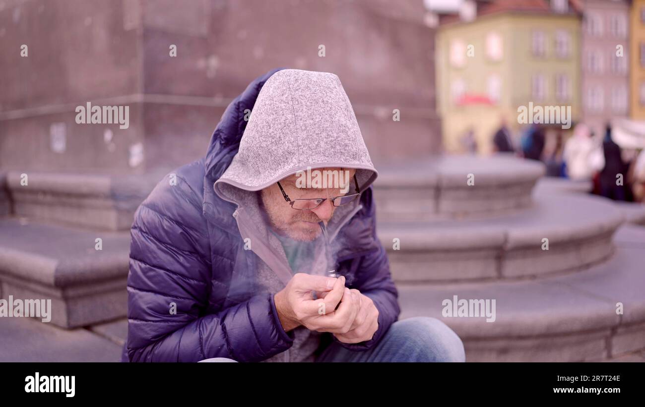 Uomo adulto Ð² Ð¾Ñ‡ÐºÐ°Ñ… Seduto sulla piazza e si illumina presso un tubo di tabacco che rilascia fumo nella Piazza del Palazzo, nella Città Vecchia di Varsavia Foto Stock