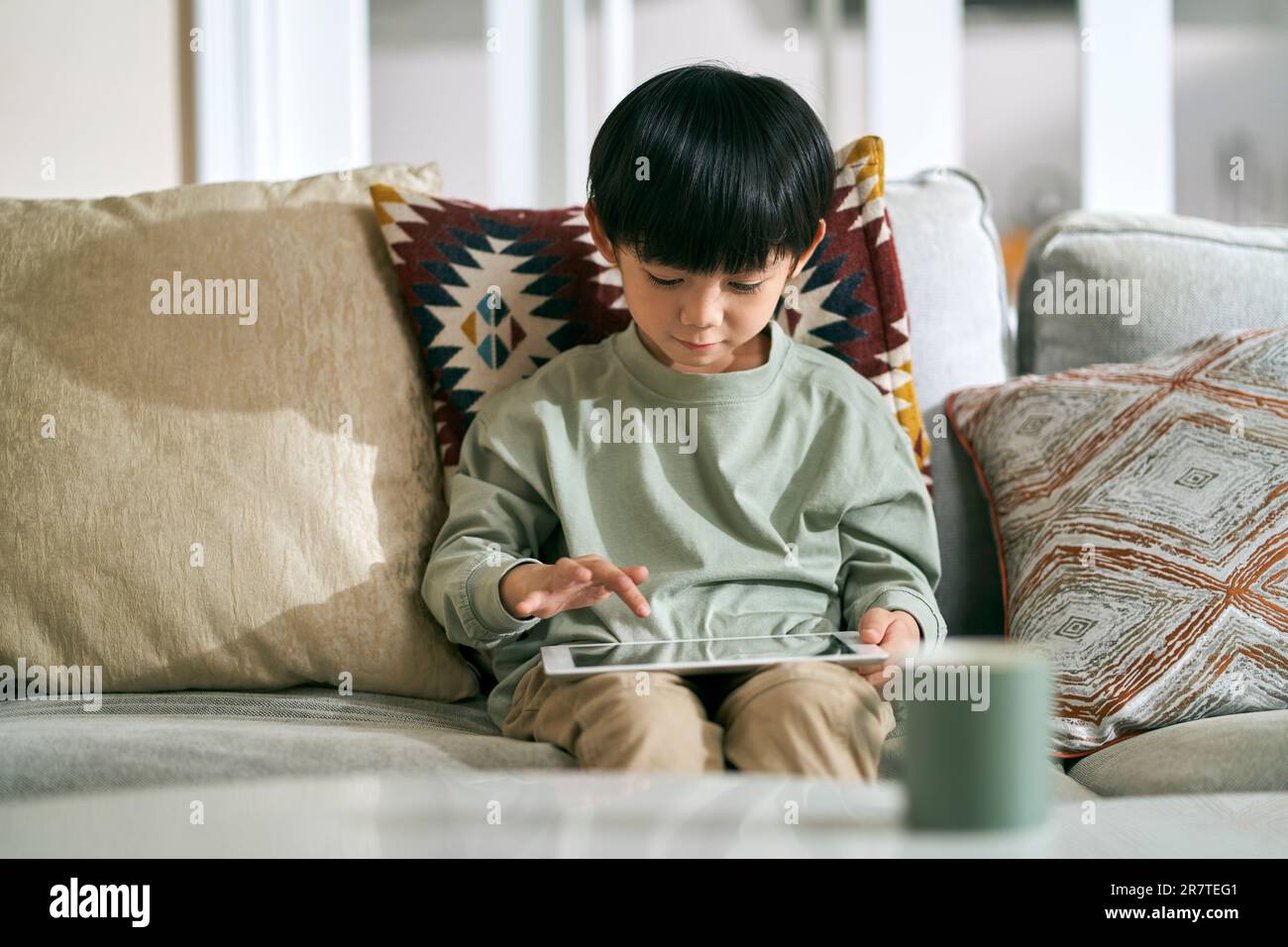 piccolo ragazzo asiatico di cinque anni seduto sul divano della famiglia a casa utilizzando un tablet digitale Foto Stock