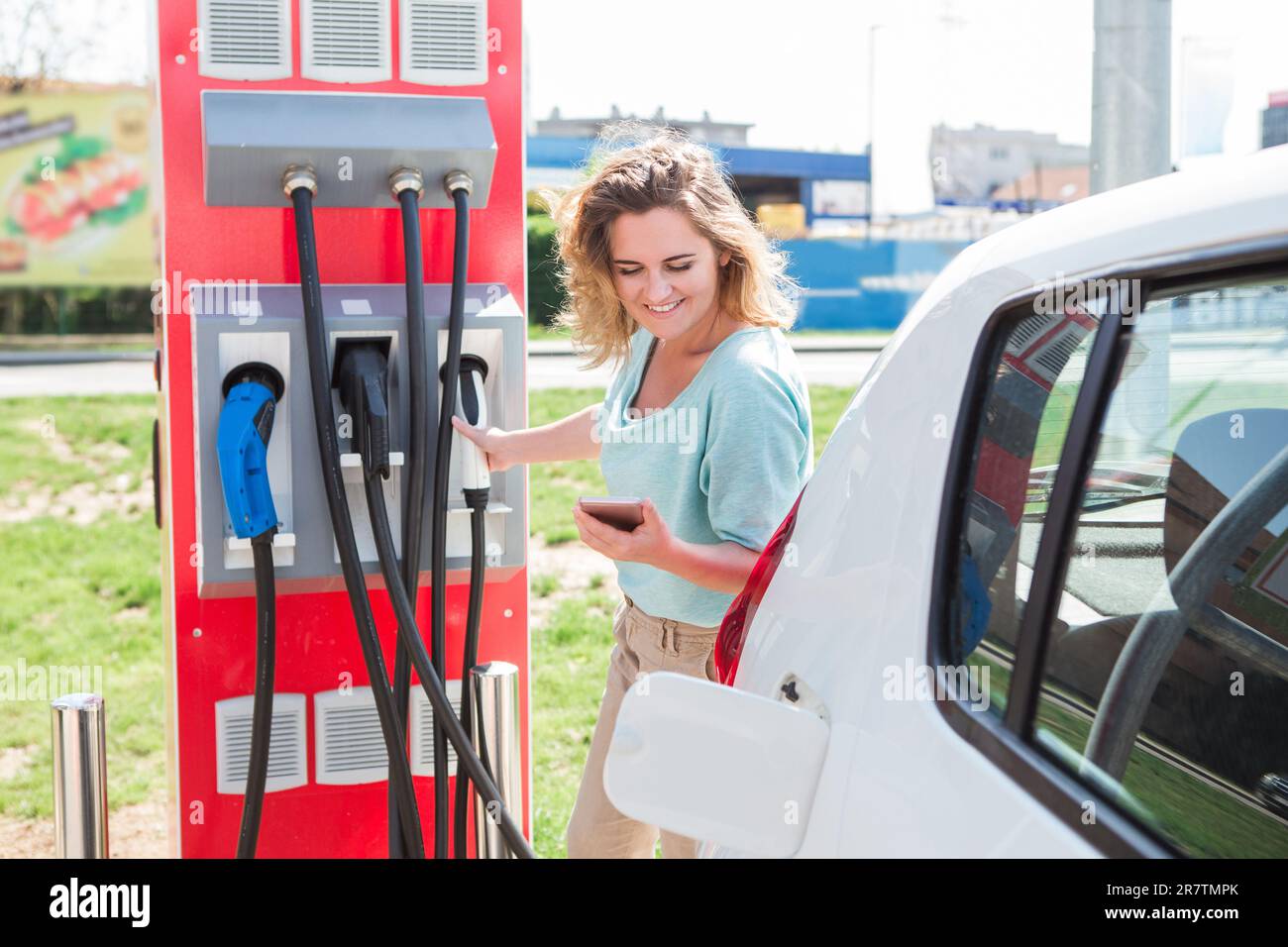 Una donna è in piedi presso la stazione di ricarica e tiene una spina del caricabatterie per un'auto elettrica. Foto Stock