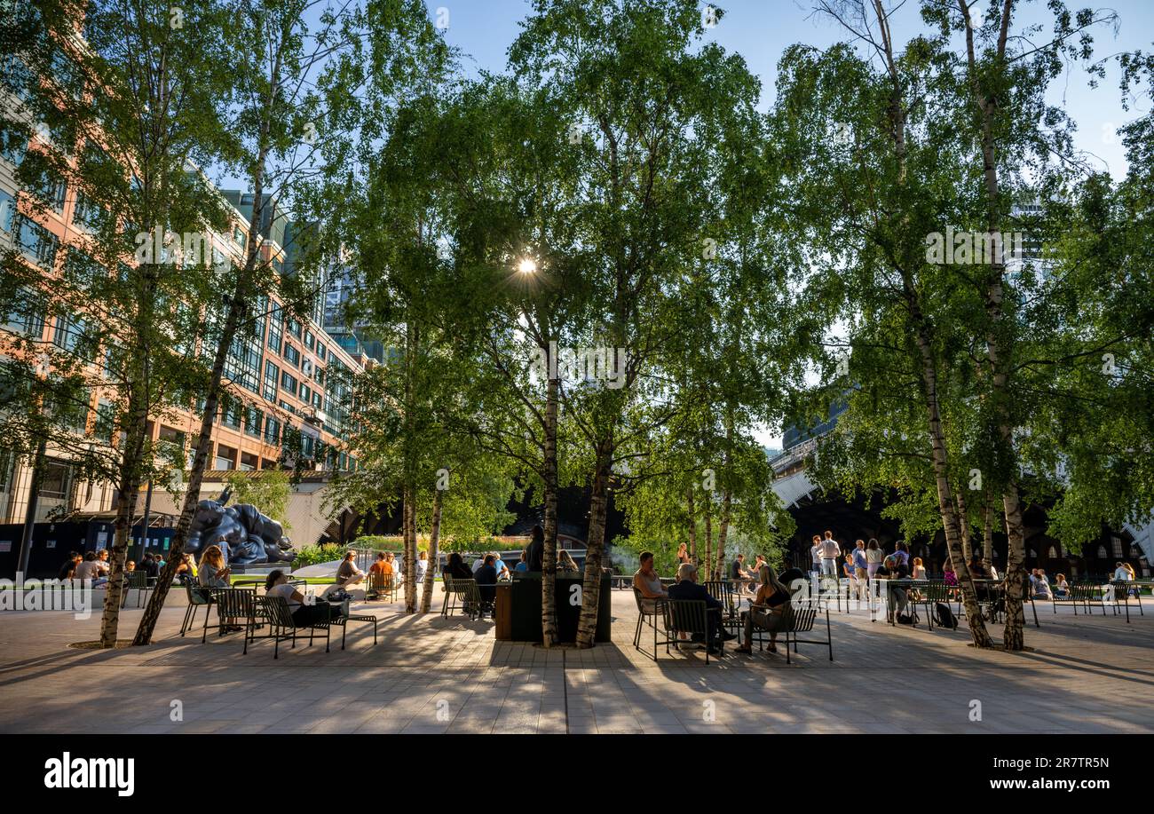 Londra, Regno Unito: Exchange Square a Broadgate, uno spazio aperto nella città di Londra. La gente si siede nella piazza e la luce del sole della sera filtra attraverso gli alberi Foto Stock