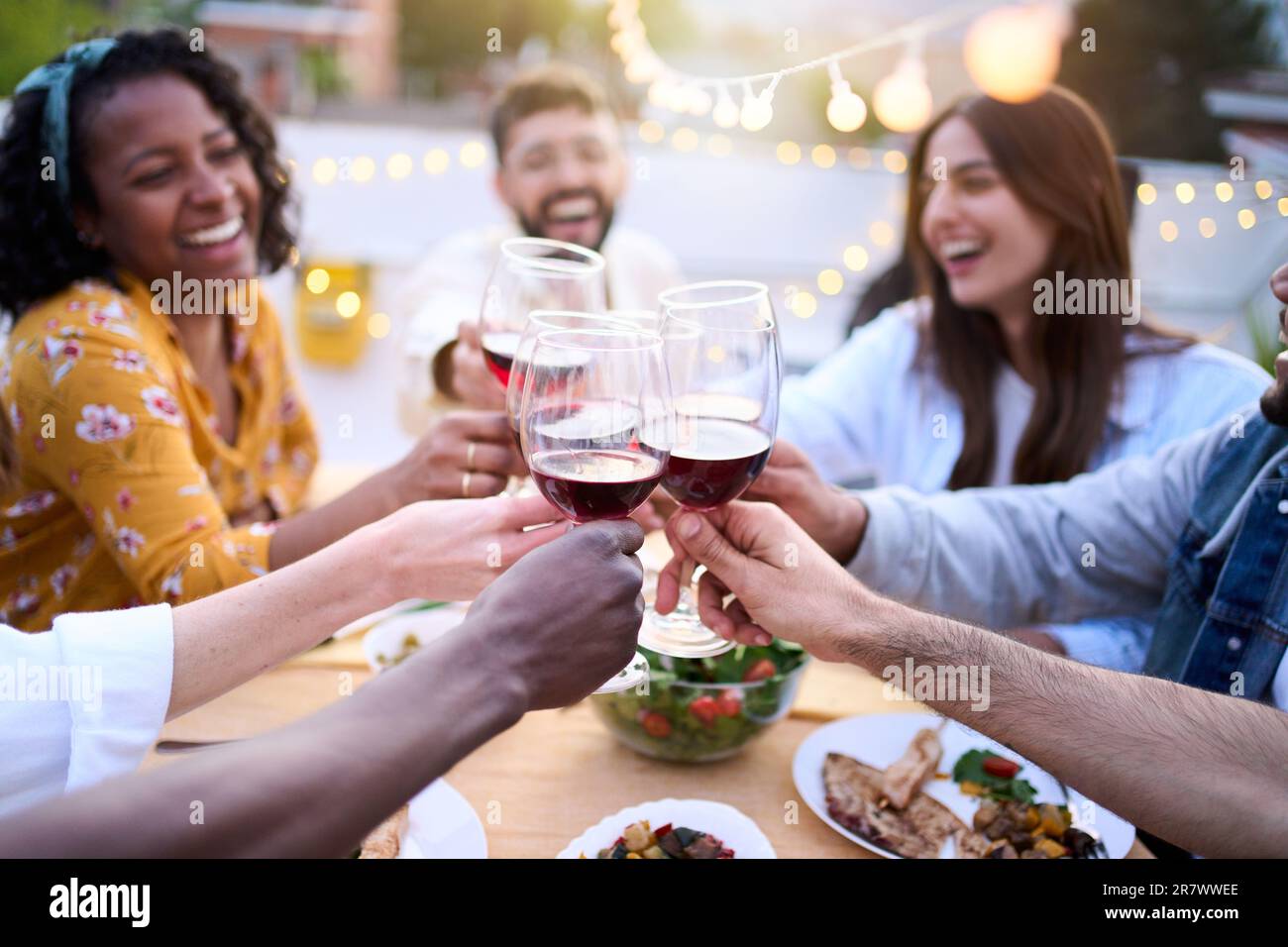 Giovani amici sorridenti che brinda vino rosso al barbecue sul tetto. Persone che bevono e mangiano all'aperto. Foto Stock