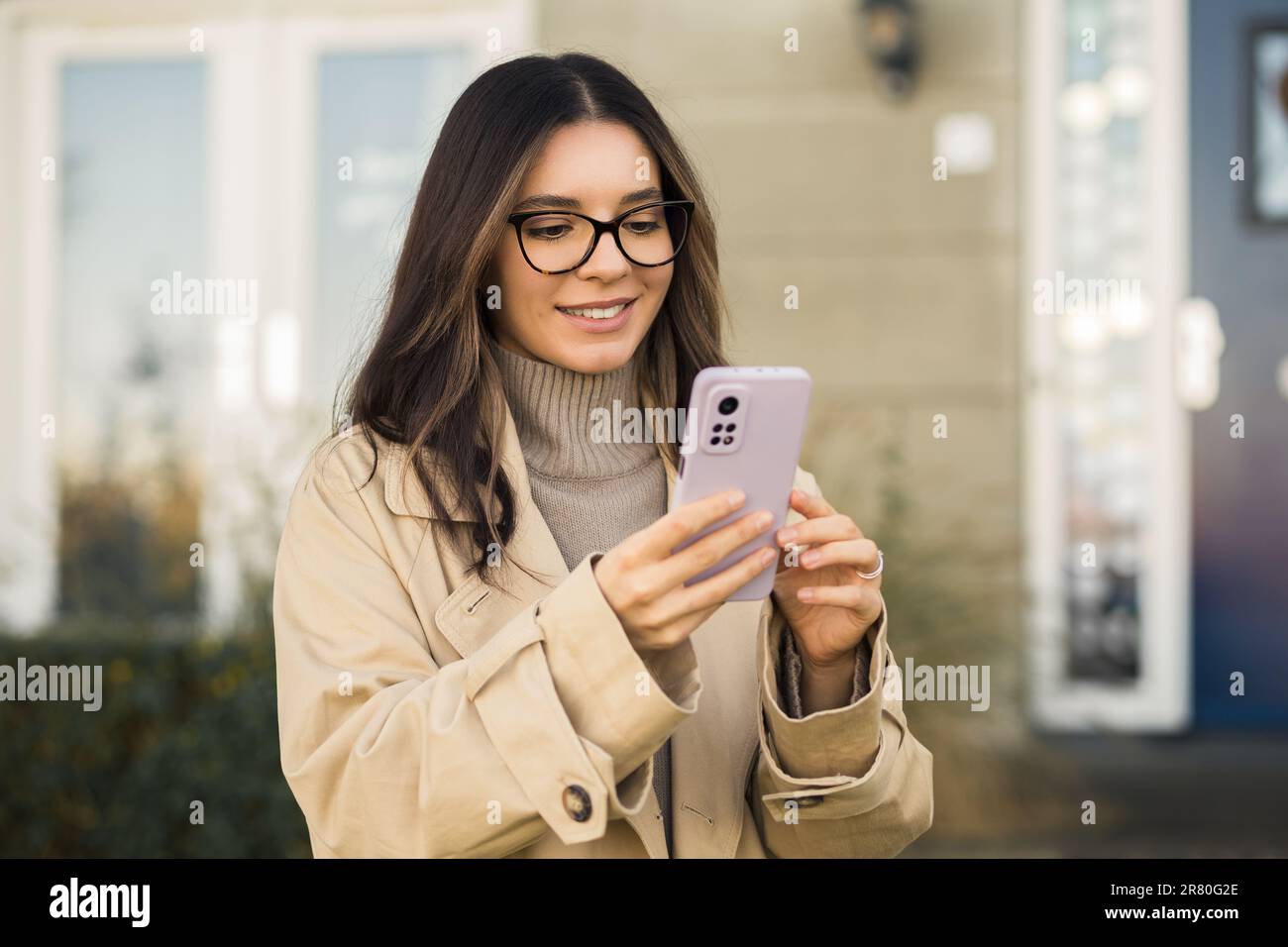 Donna sorridente in occhiali e beige trench telefono di controllo in strada di fronte al suo portico casa. Foto Stock