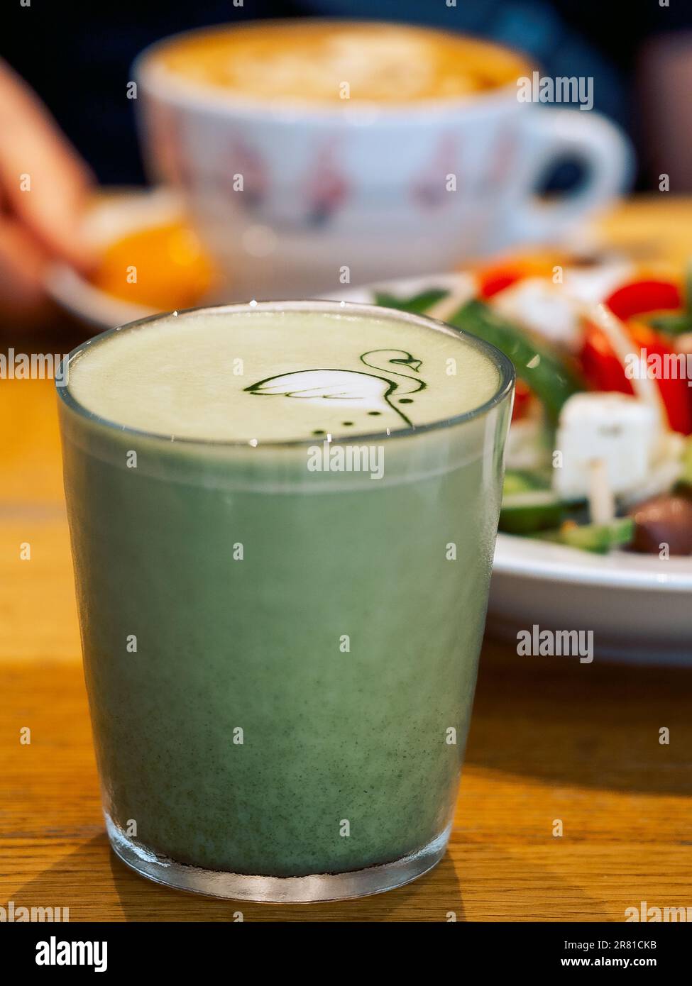 Tè matcha verde caldo in un bicchiere trasparente con arte, un piatto di insalata greca e una tazza di caffè cappuccino in un ristorante. Foto Stock