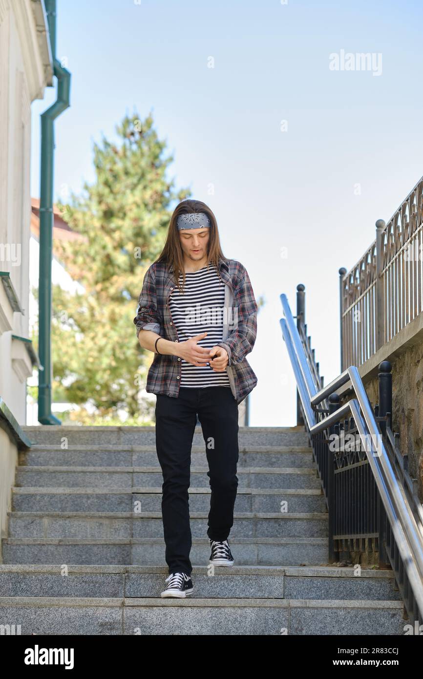 Giovane uomo dai capelli lunghi in camicia a quadri e jeans scende le scale Foto Stock