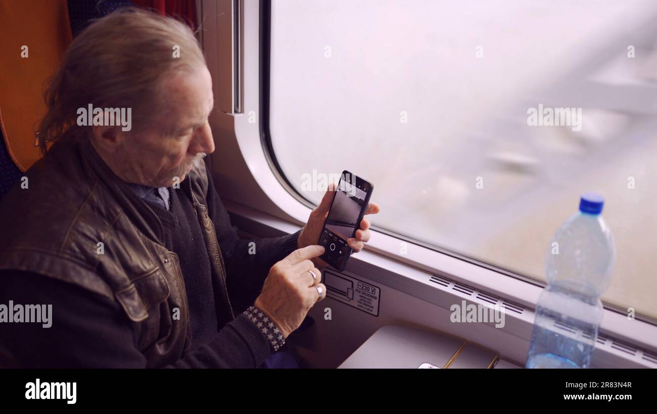 Uomo anziano che viaggia in treno e scatta foto del paesaggio attraverso la finestra utilizzando uno smartphone Foto Stock