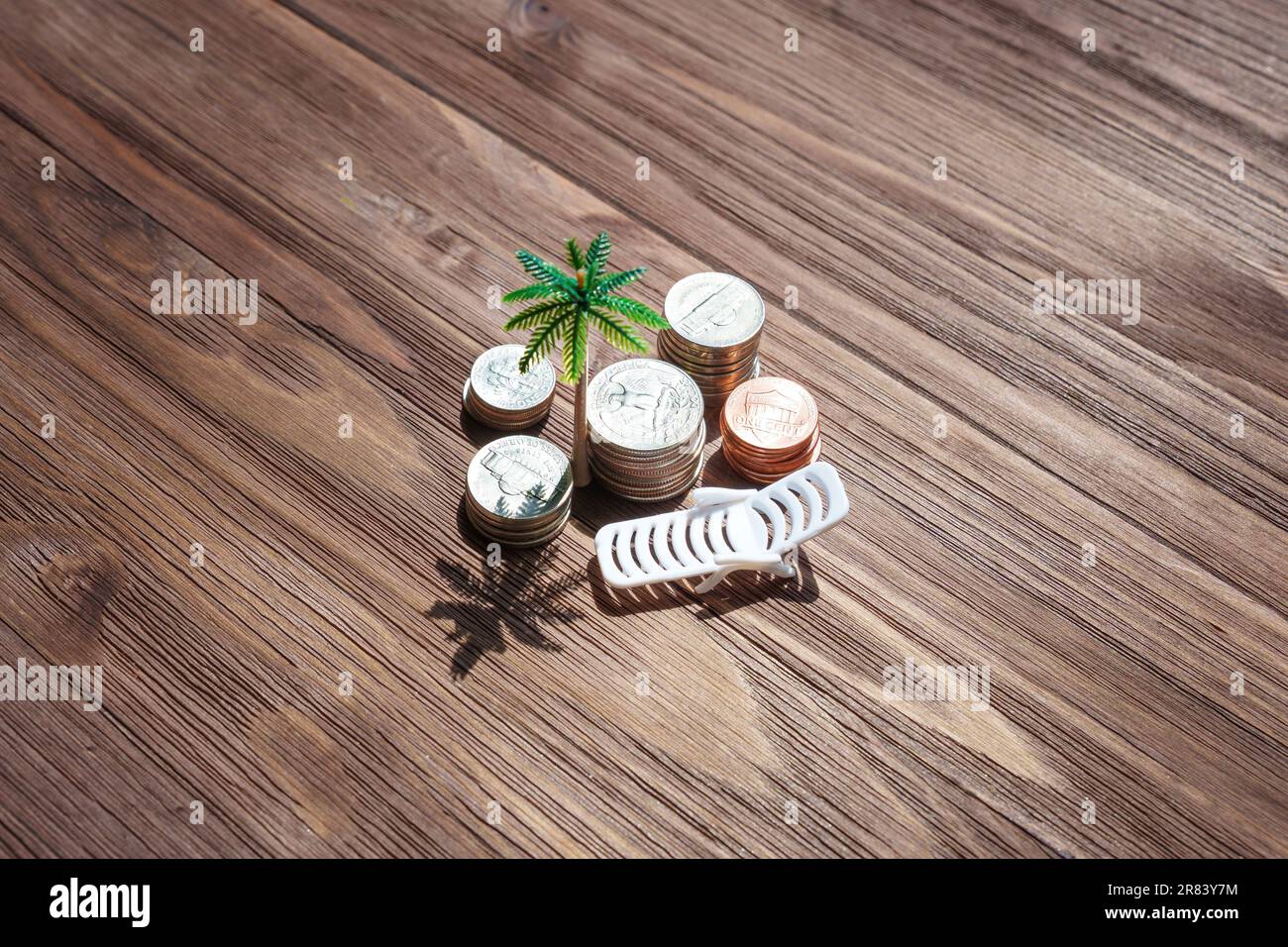 Lettino da spiaggia giocattolo e statuette di palme poste da alcune pile di monete su uno sfondo di legno scuro. Pianificazione delle vacanze, budget, obiettivi di risparmio e a. Foto Stock