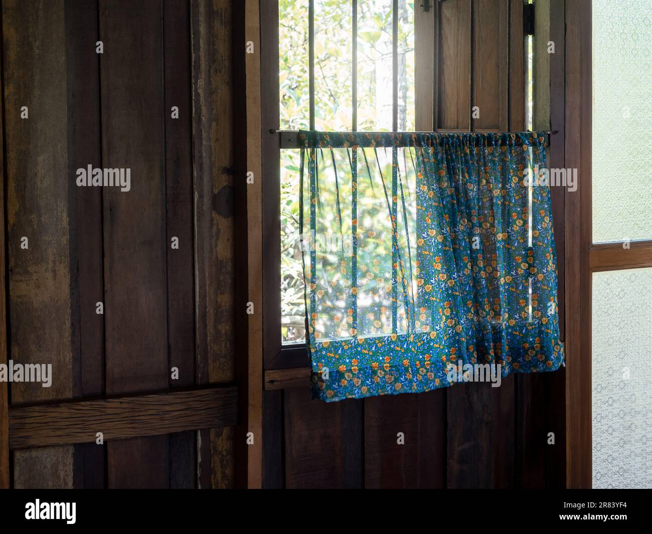 Tende floreali blu vintage su una vecchia finestra in ferro battuto in una casa di legno, leggermente soffiata dal vento dall'esterno. Una casa di campagna con un rilassato e. Foto Stock