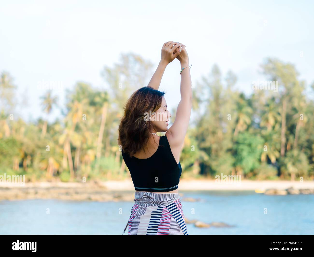 Torna della donna asiatica in nero serbatoio top chiudere gli occhi con rilassante sollevare le mani in posa sulla spiaggia in vibrazioni estive. Donna felice in piedi davanti a t Foto Stock