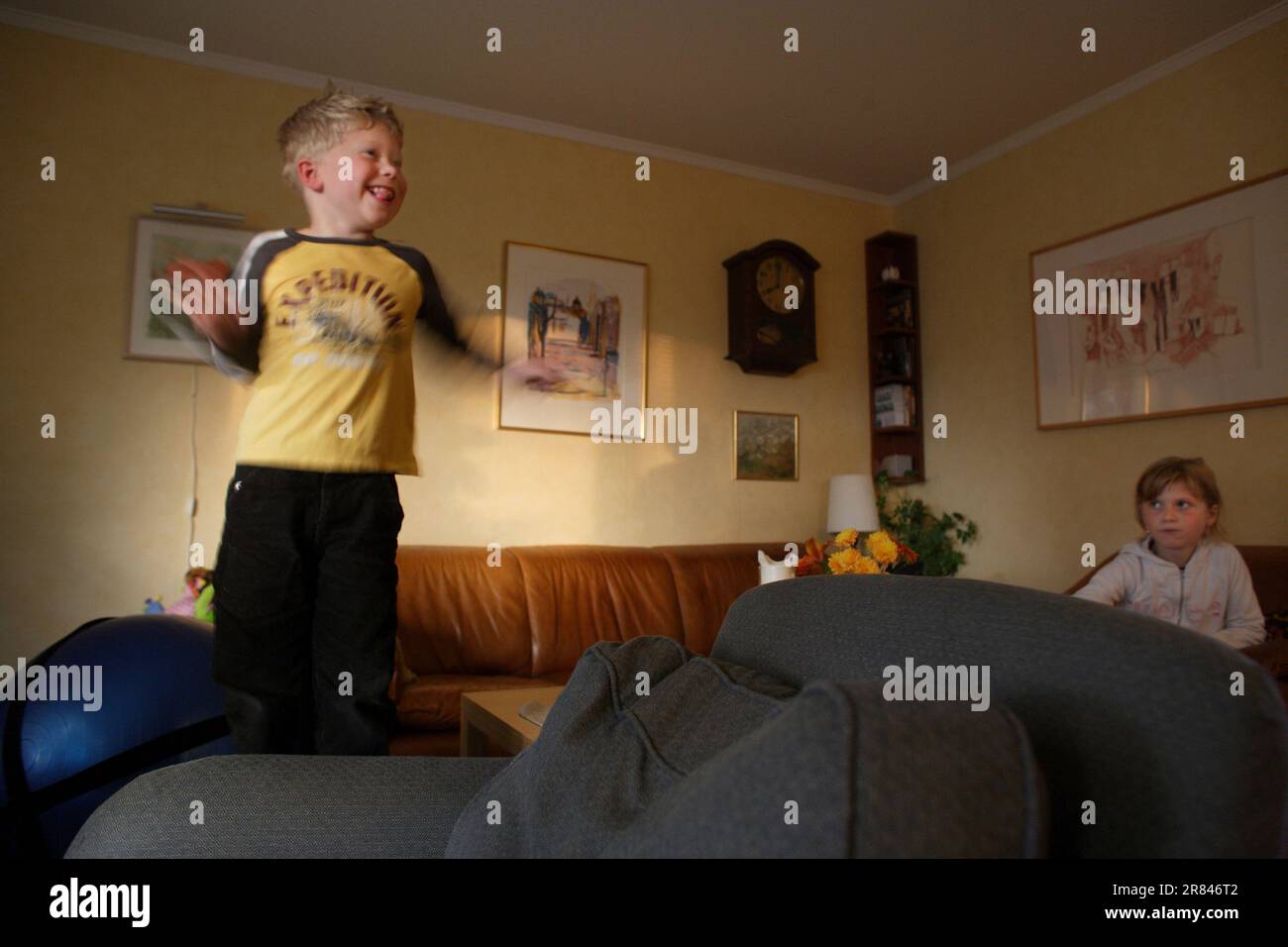 Un ragazzo autistico salta su un divano mentre la sua grande sorella lo guarda sullo sfondo. Foto Stock