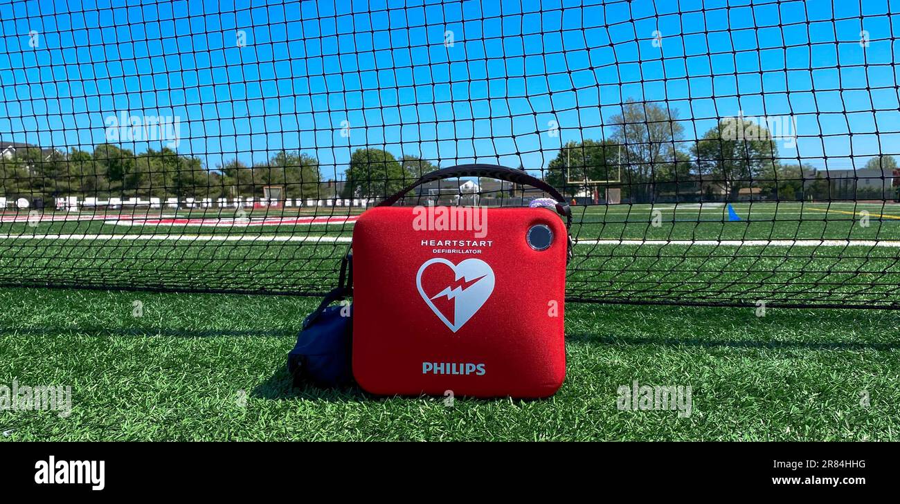 West Islip, New York, Stati Uniti d'America - 17 maggio 2023: Un defibrillatore portatile per defibrillazione semiautomatica esterna su un campo in erba davanti a una rete nera in una scuola superiore Foto Stock