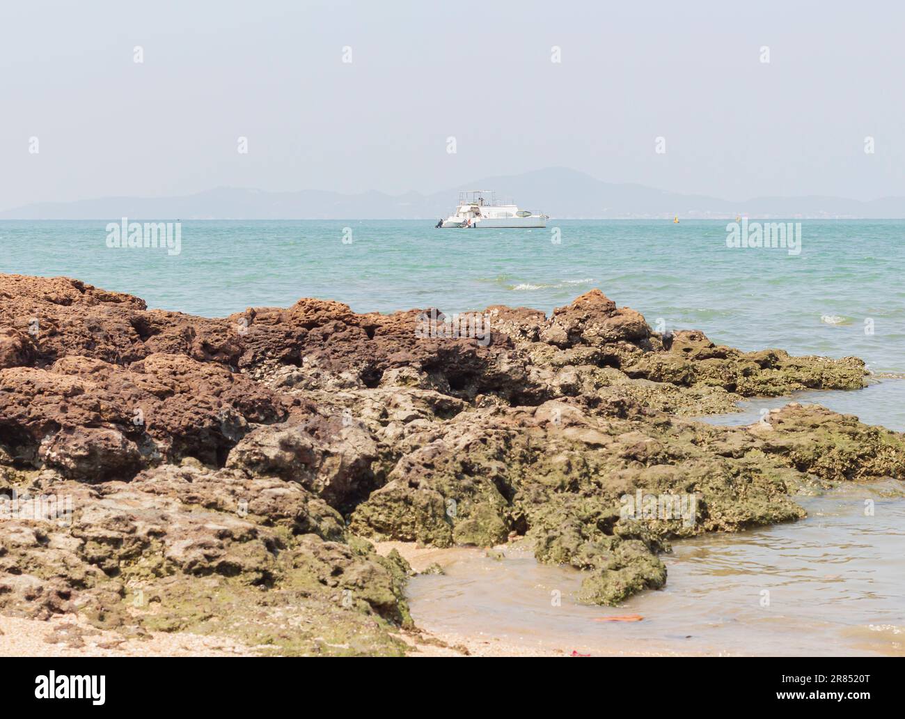 Yacht contemporaneo, sabbia, rocce in foschia, mare.Separcape. barca turistica galleggia sulle onde per l'isola. Montagne all'orizzonte. In primo piano sono grandi pietre. s Foto Stock