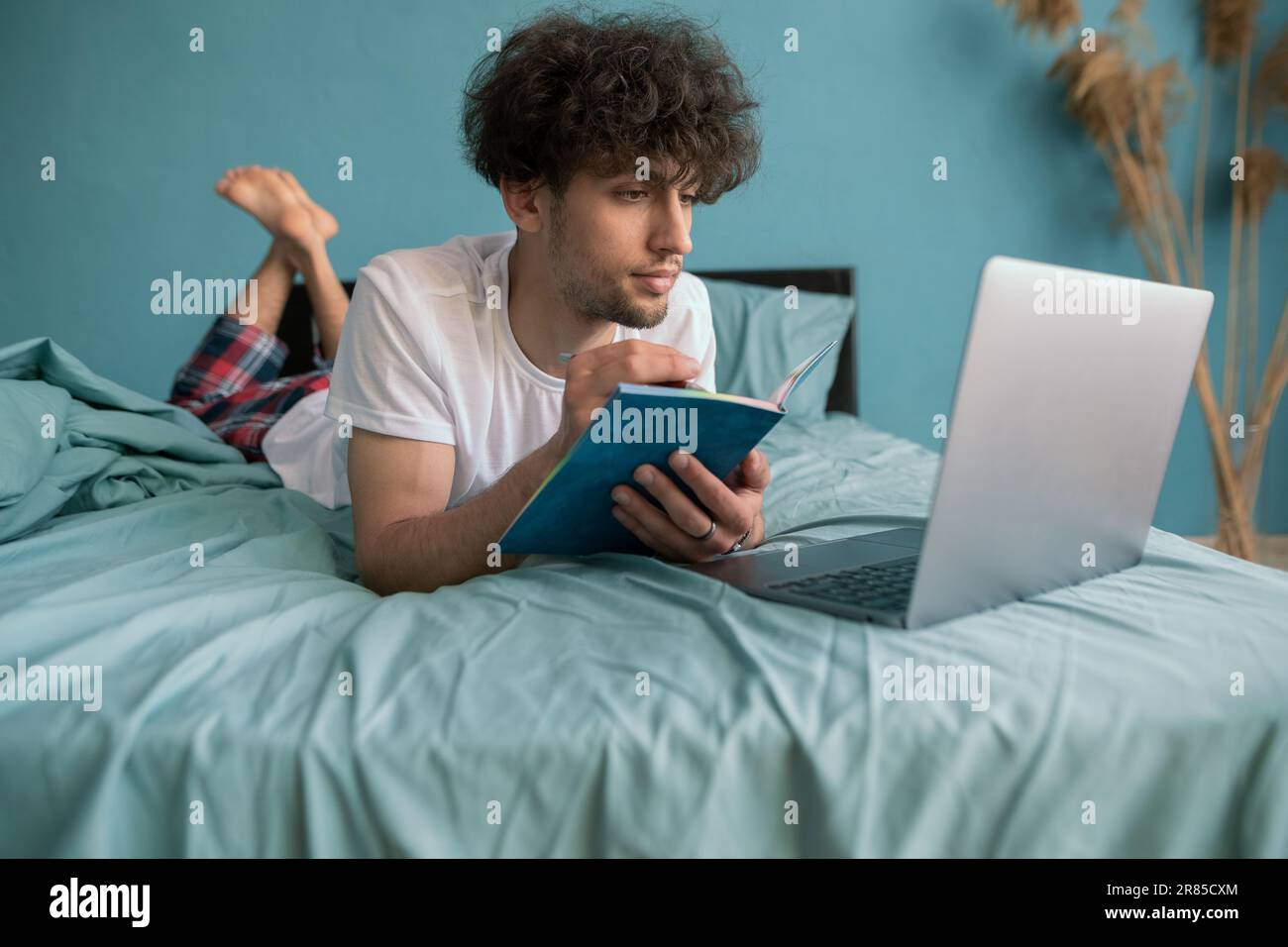 Giovane ragazzo arabo studente che studia corso virtuale, imparare a distanza prendendo formazione web, guardando il computer portatile, scrivere appunti seduti a letto Foto Stock