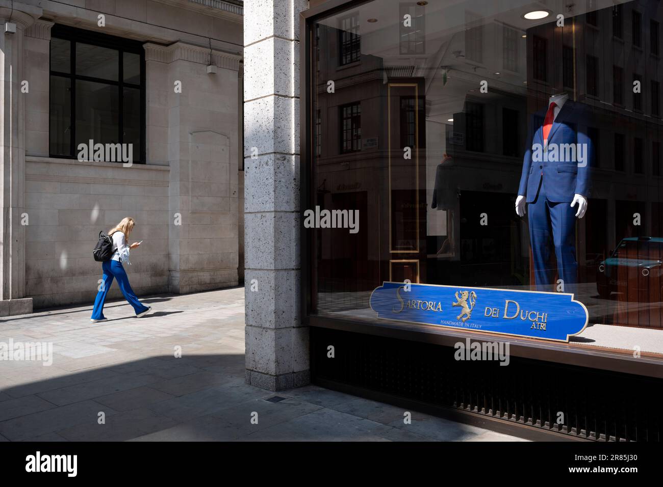 Una donna che indossa pantaloni blu passa davanti a un manichino che  indossa un abito blu coordinato di Sartoria dei Duchi su Jermyn Street nel  West End, il 19th giugno 2023, a