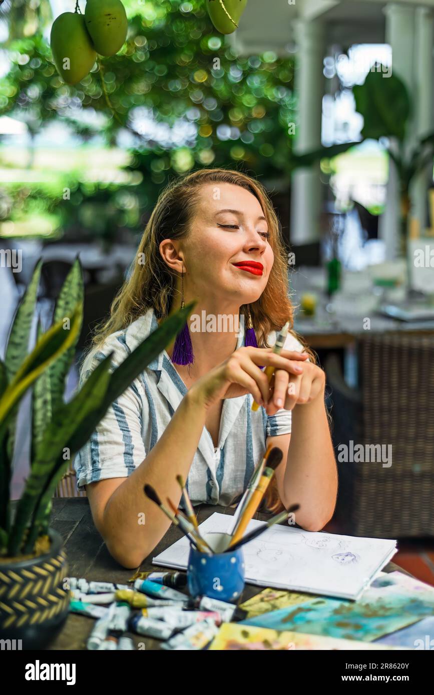 Bella artista e illustratrice bionda che pensa a un concetto del suo disegno, seduta accanto al tavolo con disegni, pennelli e pittura Foto Stock