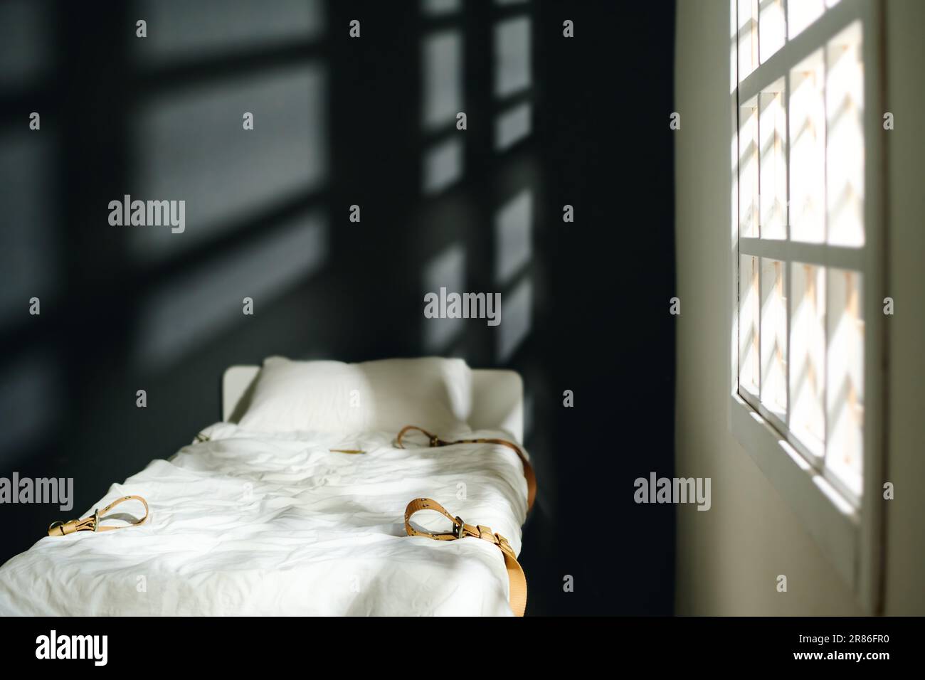Letto vuoto con cuscino e quattro cinture di pelle serrate in piedi dalla finestra con raggi di sole che penetrano nel reparto di ospedale mentale Foto Stock