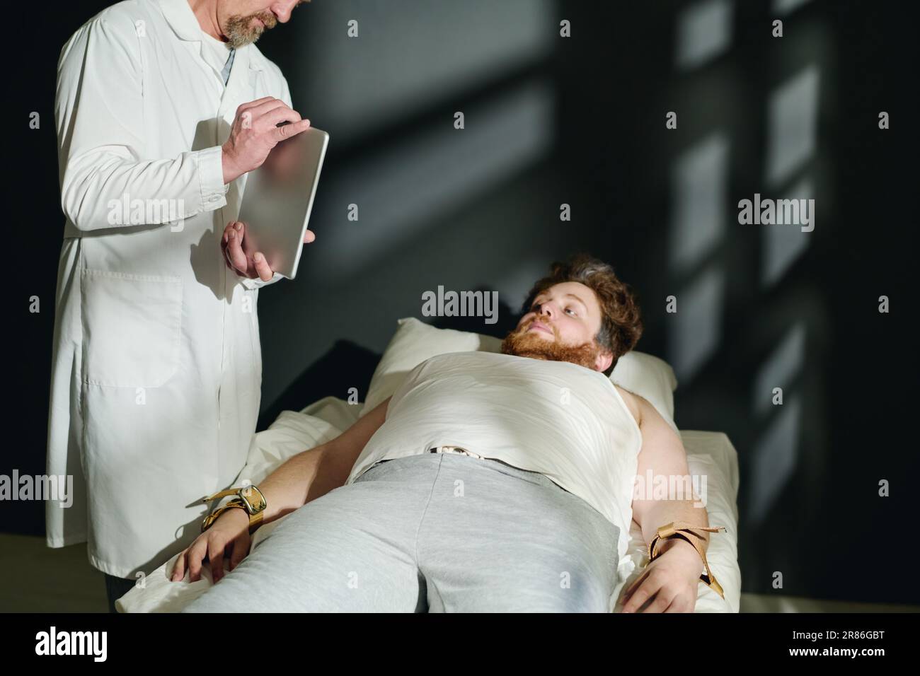 Medico maturo in camice di laboratorio che mostra qualcosa sullo schermo del tablet per il giovane paziente maschio legato a letto con cinghie strette durante il trattamento clinico Foto Stock