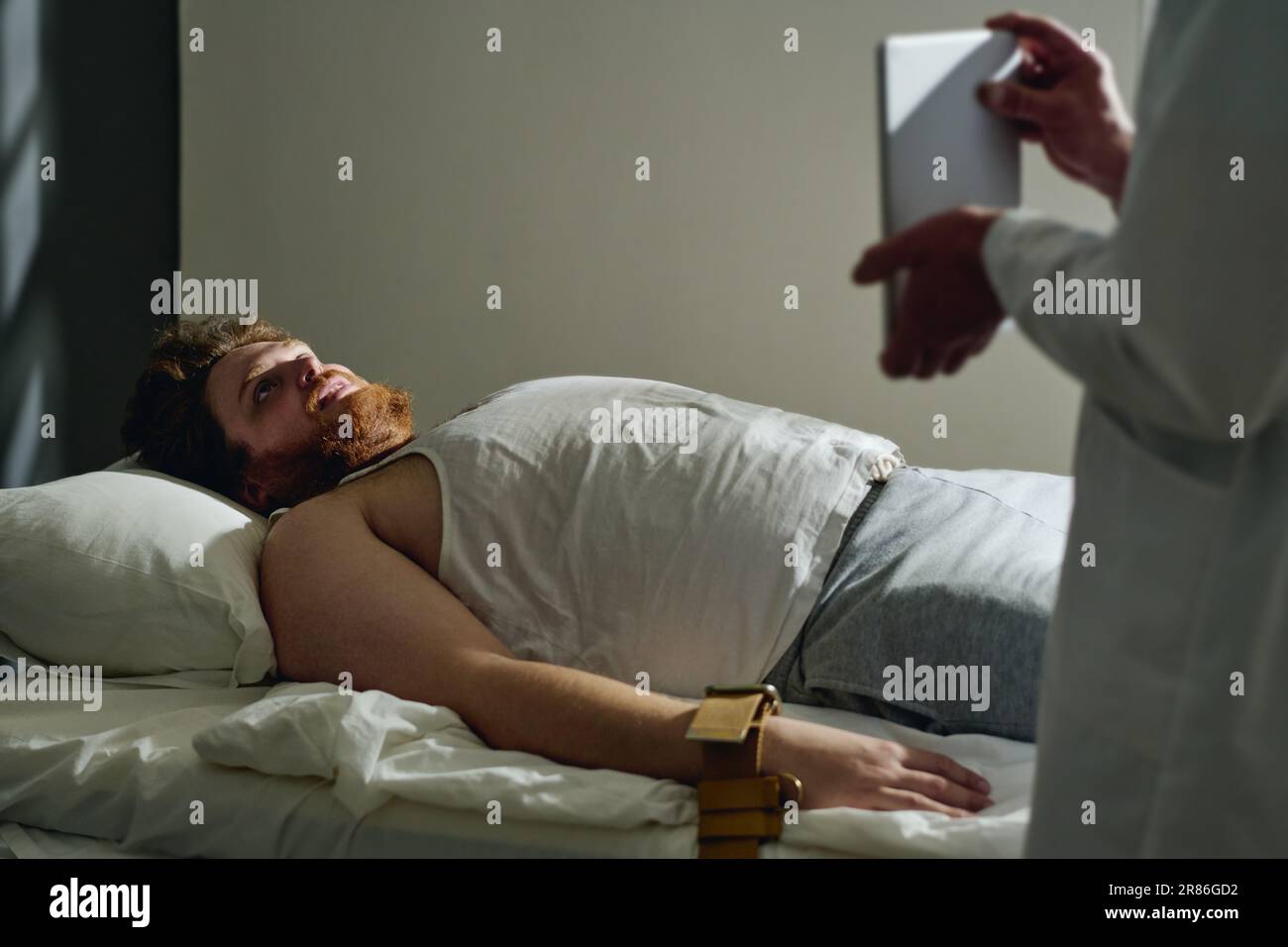 Giovane paziente maschio di ospedale mentale sdraiato nel letto con i polsi legati con le cinture strette e guardando lo schermo della compressa tenuto dal medico Foto Stock