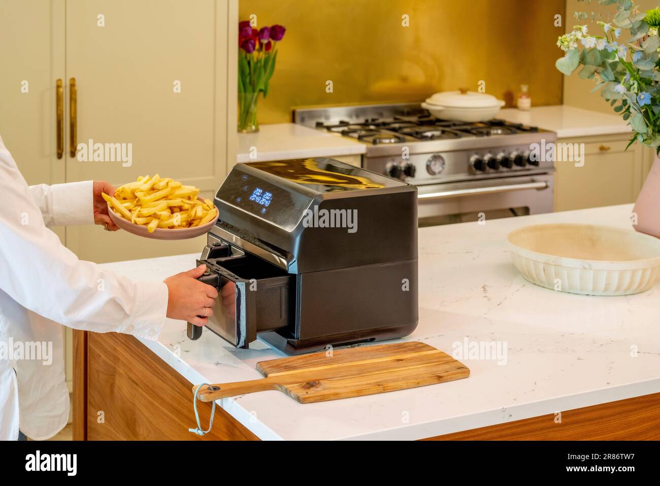 Mani che tengono un piatto di patatine crude mentre si apre un cassetto su una friggitrice ad aria, in una cucina moderna con un tradizionale fornello ranger in lontananza. Foto Stock