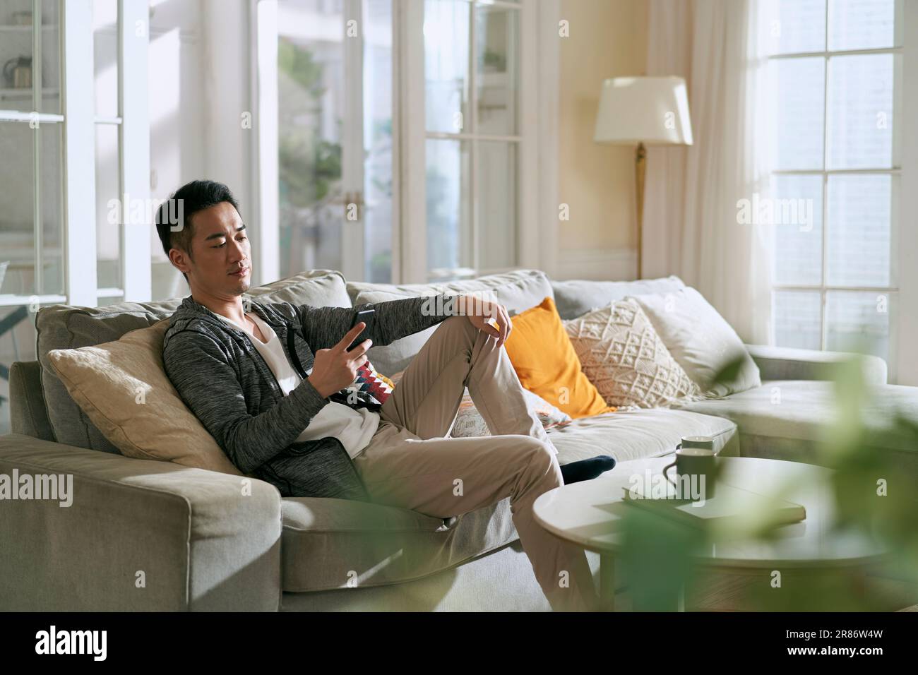 giovane uomo asiatico adulto seduto sul divano della famiglia nel soggiorno di casa guardando il cellulare Foto Stock