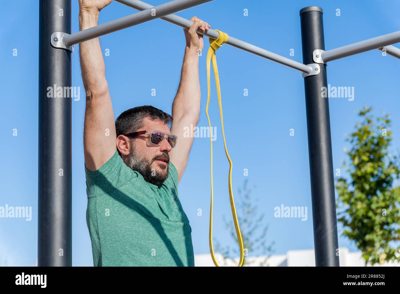 Uomo bearded con occhiali da sole visto in profilo allenare la schiena facendo pull-up su un barbell con una fascia elastica fitness in una palestra all'aperto Foto Stock