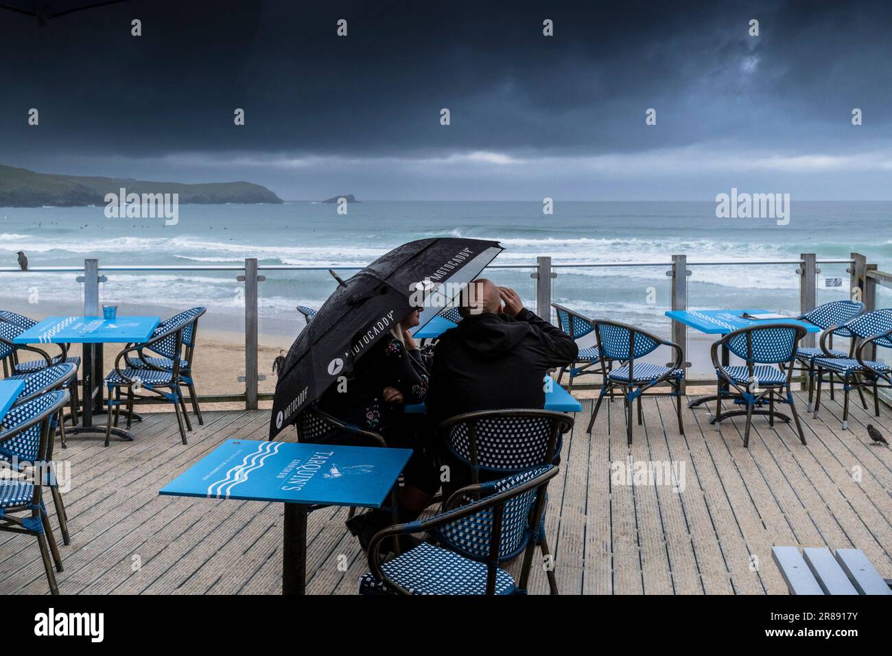 Meteo nel Regno Unito. Una coppia che si ripara sotto un ombrellone seduto ad un tavolo sulla terrazza esterna del Fistral Beach Bar che si affaccia sulla spiaggia di Fistral Foto Stock