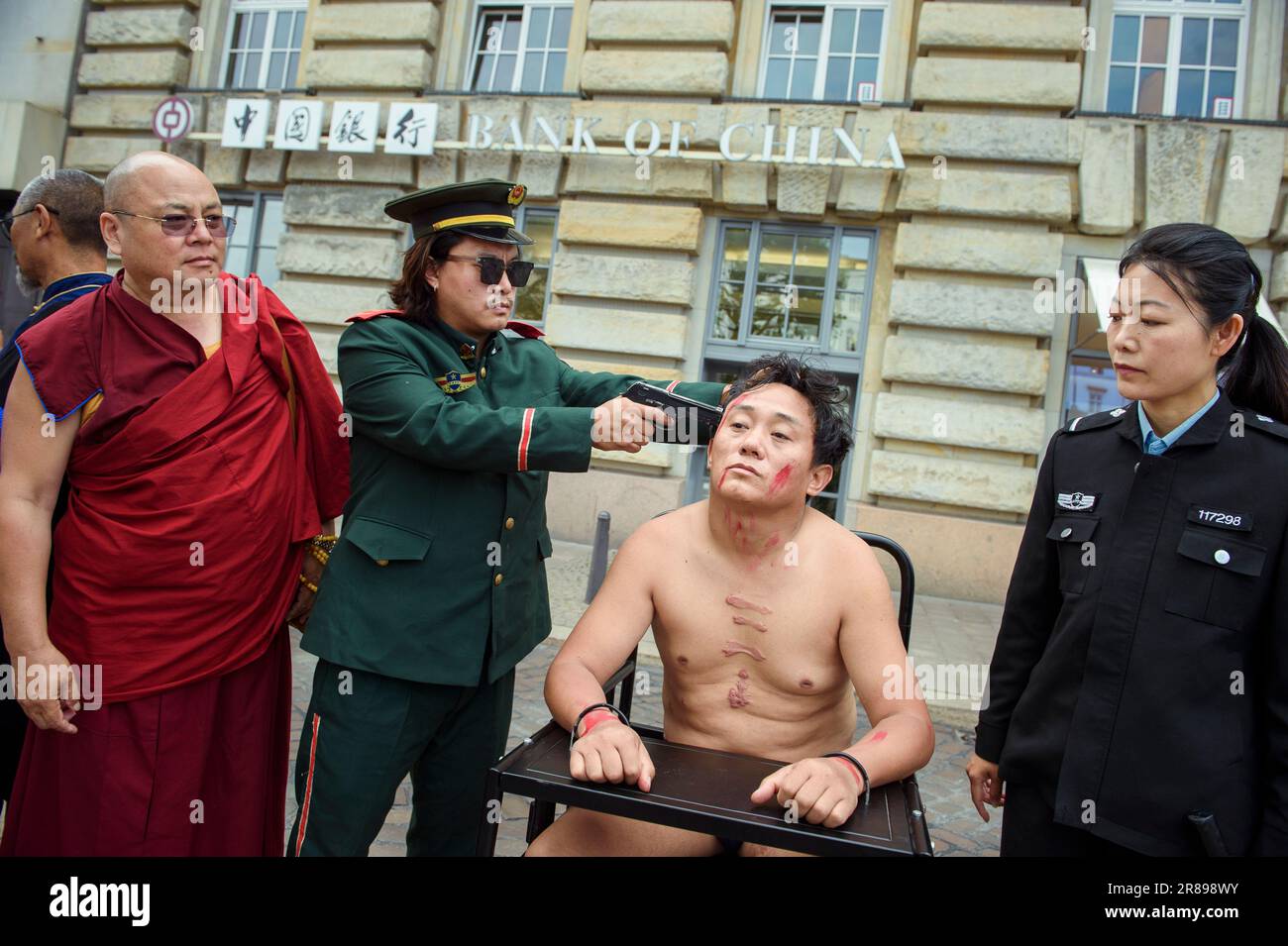 Amburgo, Germania. 20th giugno, 2023. Golog Jigme Gyatso (l), attivista per i diritti umani e regista, partecipa a un'azione di protesta da parte di attori tibetani contro i crimini sui diritti umani in Tibet, Mentre un attivista siede su un'imitazione di una sedia tigre di fronte ad un ramo della Banca della Cina al Rathausmarkt ed è minacciato con un'arma fittizia. Su una cosiddetta sedia della tigre, le persone sono immobilizzate durante l'interrogazione. Credit: Gregor Fischer/dpa/Alamy Live News Foto Stock