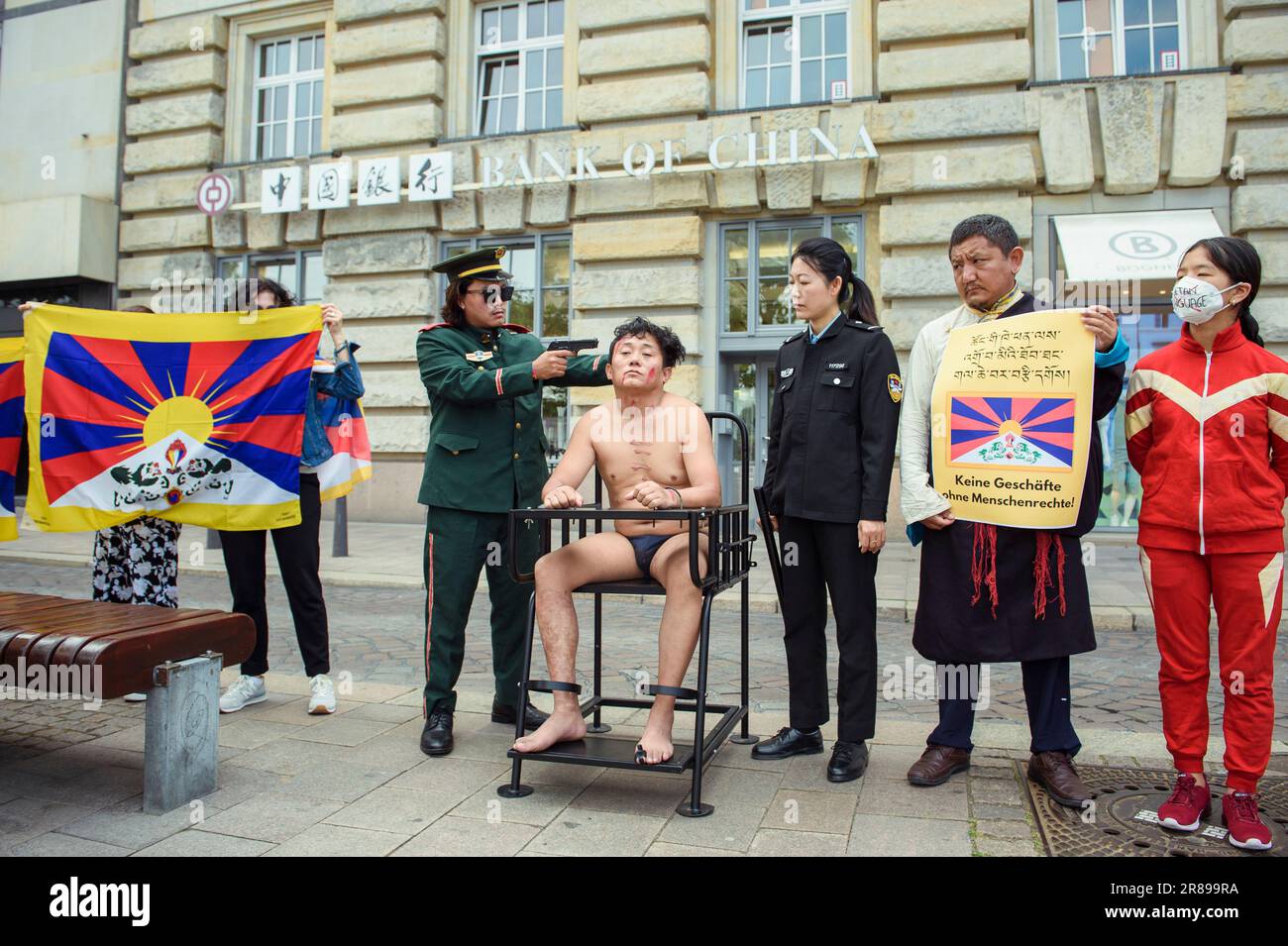 Amburgo, Germania. 20th giugno, 2023. Un attivista (M) siede su un'imitazione di una sedia di tigre durante un'azione di protesta degli attori tibetani contro i crimini dei diritti umani in Tibet di fronte a un ramo della Banca della Cina presso il Rathausmarkt ed è minacciato da un'arma fittizia. Su una cosiddetta sedia della tigre, le persone sono immobilizzate durante l'interrogazione. Credit: Gregor Fischer/dpa/Alamy Live News Foto Stock