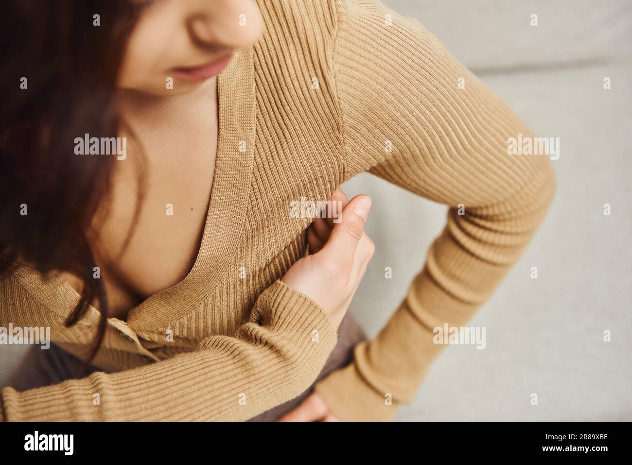Vista dall'alto di una giovane bruna in saltatore marrone che massaggia i nodi linfatici sulle ascelle durante il massaggio di drenaggio sul divano a casa, migliorando l'autoapprendimento Foto Stock