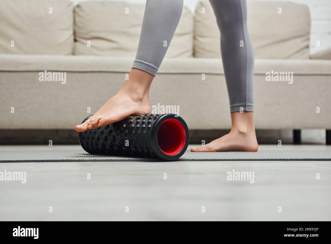 Vista ritagliata della donna a piedi nudi in scarpe sportive con massaggiatore a rulli e in piedi sul tappetino fitness vicino al divano a casa, per favorire il flusso linfatico Foto Stock