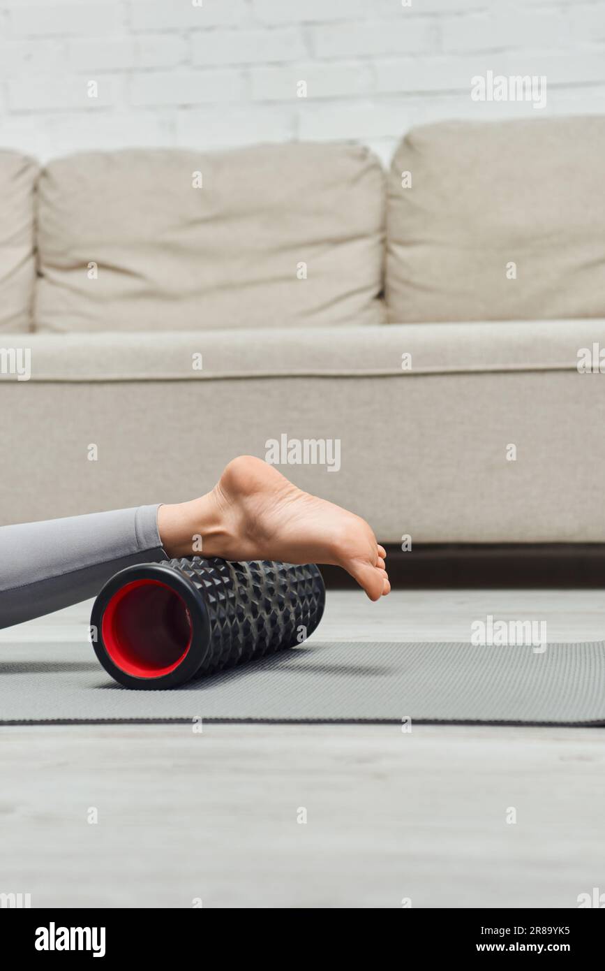 Vista ritagliata della donna a piedi nudi che massaggia le gambe con un moderno massaggiatore a rulli mentre si sdraiava sul tappetino fitness vicino al soggiorno di casa, favorendo il flusso linfatico e. Foto Stock
