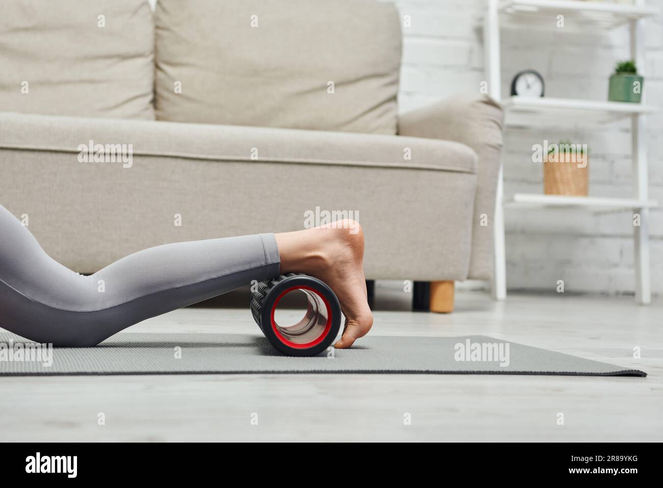 Vista ritagliata della donna a piedi nudi in abbigliamento sportivo gambe massaggianti con massaggiatore a rulli sul tappetino fitness vicino al divano nel soggiorno di casa, promuovendo il flo linfatico Foto Stock