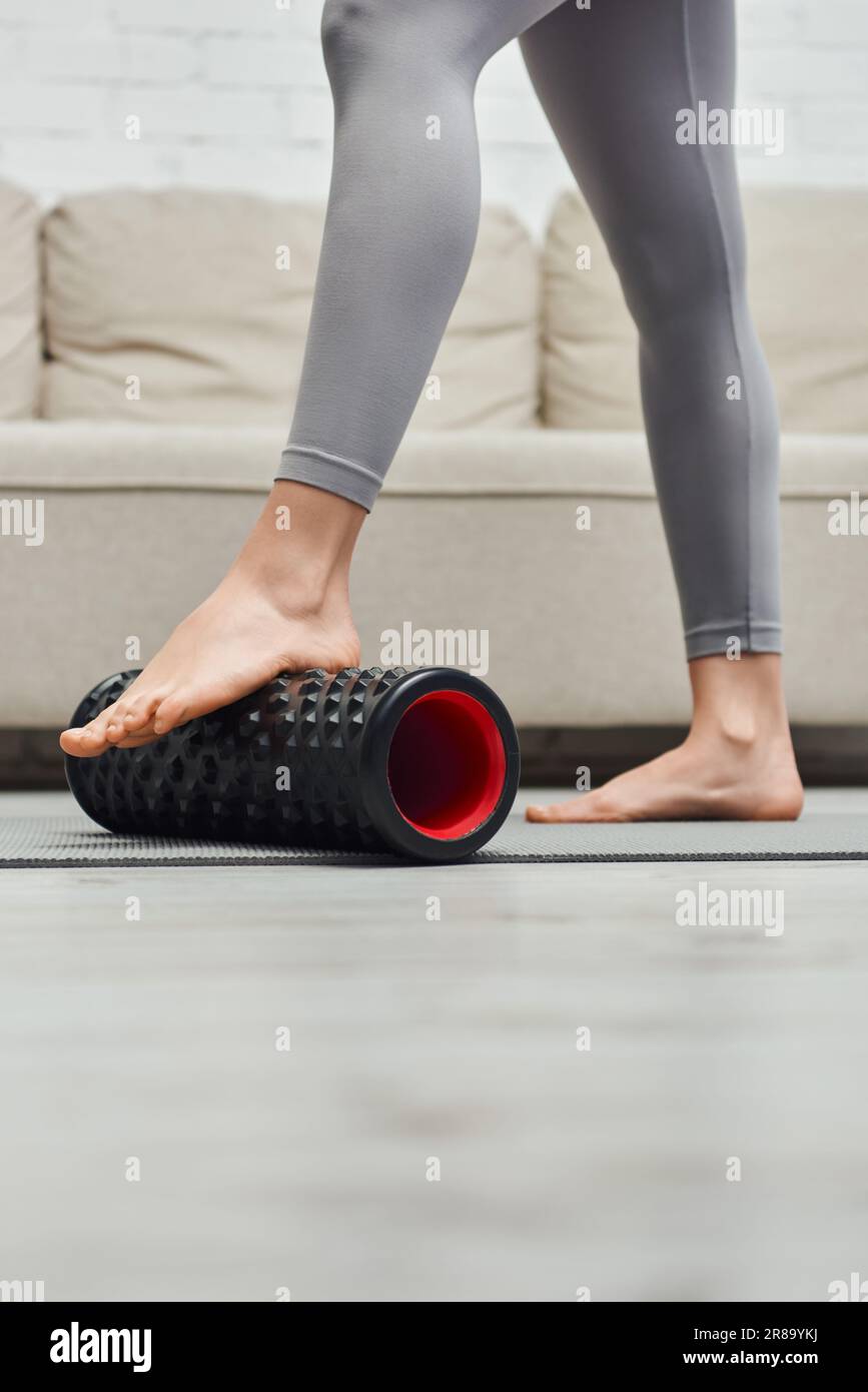 Vista ritagliata della donna a piedi nudi in piedi sportivi con massaggiatore a rulli moderno sul tappetino fitness vicino al divano nel soggiorno, per favorire il flusso linfatico Foto Stock