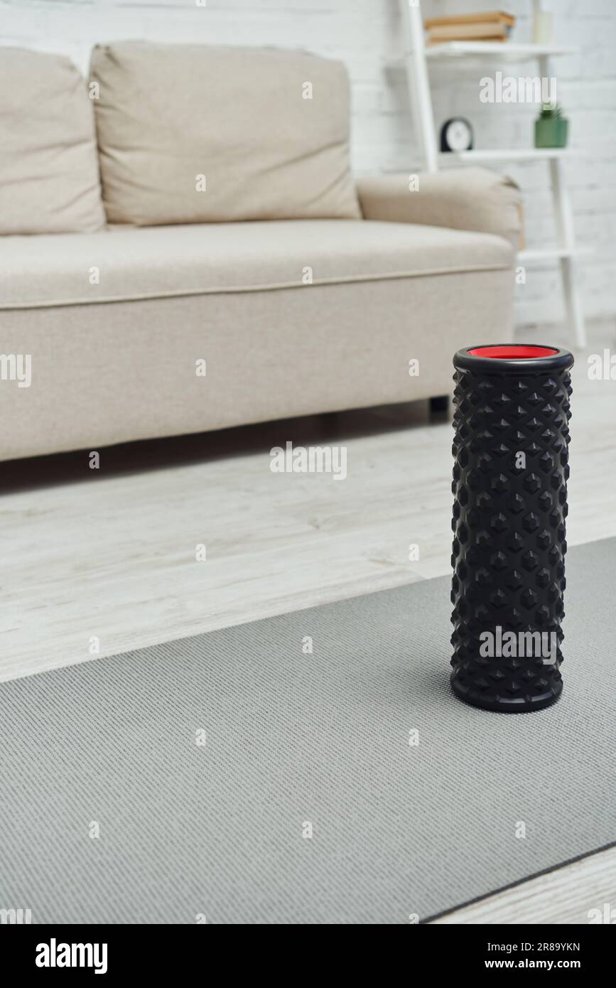 Moderno massaggiatore a rulli nero in piedi sul tappetino fitness vicino al divano sfocato nel soggiorno di casa, promuovendo il flusso linfatico e il benessere a casa Foto Stock