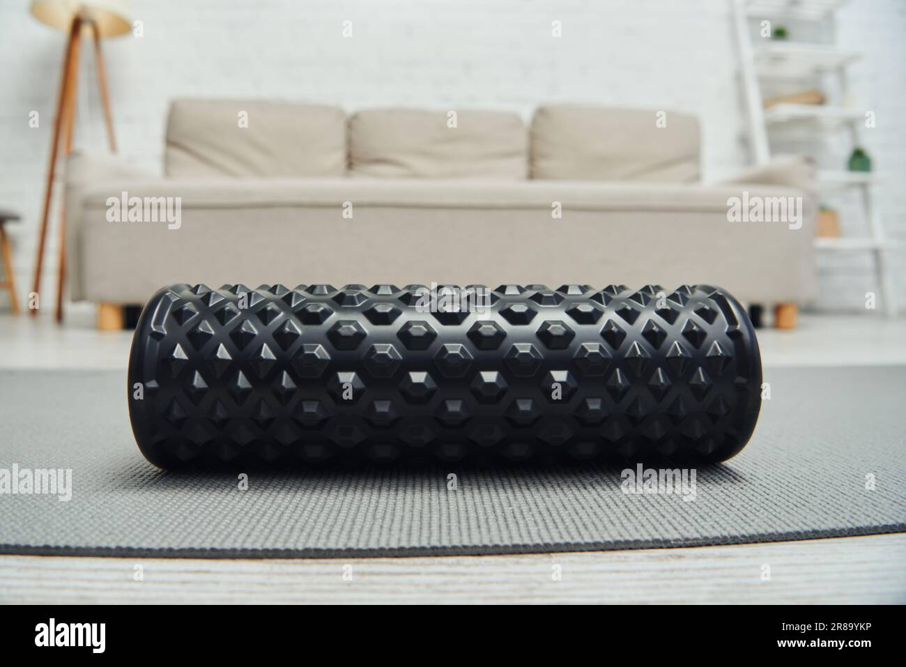 Vista ravvicinata del massaggiatore a rulli nero sdraiato sul tappetino fitness vicino al divano sfocato nel soggiorno di casa, favorendo il flusso linfatico e il benessere della casa Foto Stock
