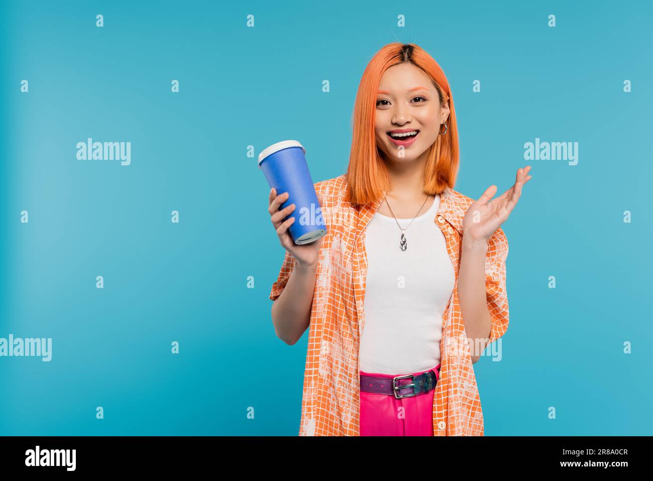 positività, bere in mano, felice asiatico e giovane donna con capelli rossi tenendo la tazza di carta e guardando la macchina fotografica su sfondo blu, abbigliamento casual, generi Foto Stock