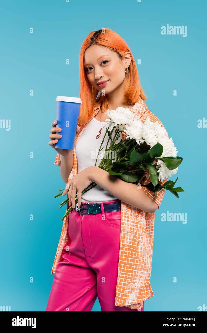 vibrazioni primaverili, asiatica e giovane donna con tinto capelli rossi che tiene la tazza di carta e bouquet di fiori su sfondo blu, abbigliamento casual, generazione z, takea Foto Stock