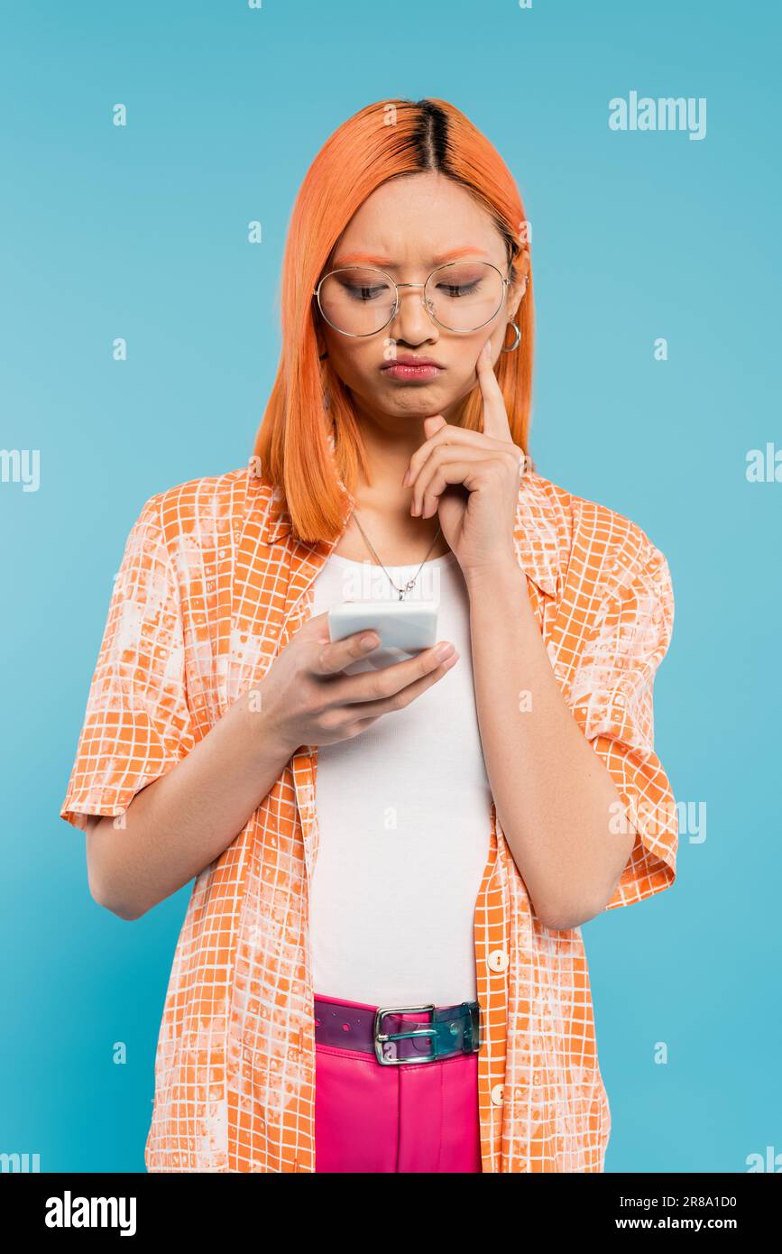 emozione negativa, cattivo umore, donna asiatica dispiaciuta toccare guancia mentre si guarda lo smartphone su sfondo blu, occhiali alla moda, capelli rossi, Foto Stock